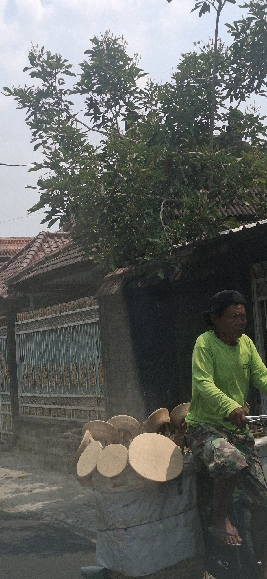 The image depicts a man riding a bicycle loaded with circular wooden objects. He is wearing a green long-sleeved shirt and camouflage pants, and a black cap. The bicycle is laden with numerous round, flat wooden pieces, some stacked on top of others, and appears to be covered with a grey tarp at the bottom. The man is on a paved surface, possibly a street, with a weathered building and a decorative metal fence in the background. Lush green foliage from a large tree overhangs the scene from above. The sky is overcast. The location is identified as Gajahan, Indonesia.