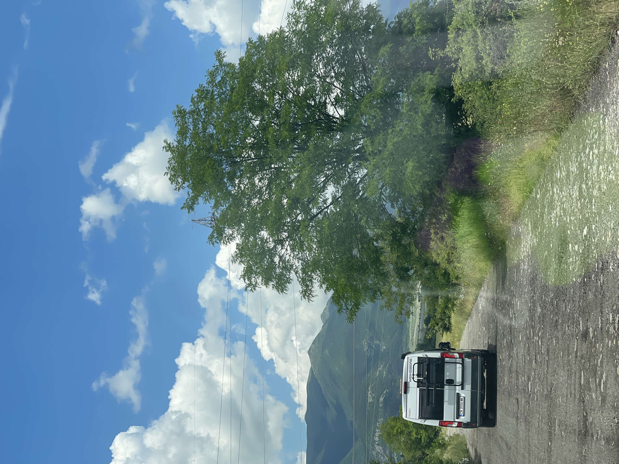 A white Weinsberg CaraBus camper van is observed from a rear perspective, positioned on an unpaved, gravel-strewn dirt road in what is identified as Dunavtsi, Bulgaria. The vehicle displays a Bulgarian license plate, "CB 3556 MK," and features a black bicycle rack mounted on its rear doors, though no bicycles are present.
The road extends into the background, flanked by overgrown green grass and various types of foliage and low-lying shrubs. To the immediate right, a large, dense-canopied tree dominates the mid-ground. In the distance, multiple layers of green, undulating mountains rise under a bright blue sky filled with scattered white cumulus clouds. Several parallel power lines traverse the middle-ground, with a section of a metallic power transmission tower partially visible on the far left. The scene is illuminated by natural daylight, suggesting a clear day. No individuals are visible in the image.