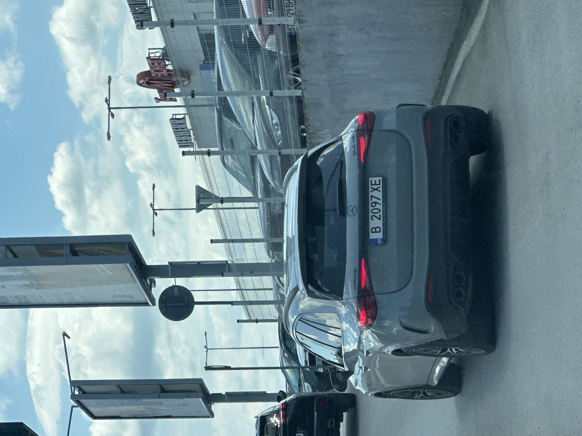 The image captures a rear view of a gray Mercedes-Benz wagon, identifiable by the "GLC 4S" badging on its tailgate. The car displays a Bulgarian license plate, "B 2097 XE", with "BG" visible to the left of the plate. The car is situated in a lane of traffic, with other vehicles visible in the background, including a black SUV to the left. Behind the vehicles, a concrete wall and a metal fence separate the roadway from a commercial area with buildings and signage. One sign reads "METRO" in large red letters, and another displays "LIEBHERR". The sky is partly cloudy with a bright, overcast atmosphere. The scene suggests an urban or suburban environment, likely a commercial district with a main road. The specific location in Veliko Tarnovo, Bulgaria, is implied by the license plate, though no explicit landmarks confirm this.