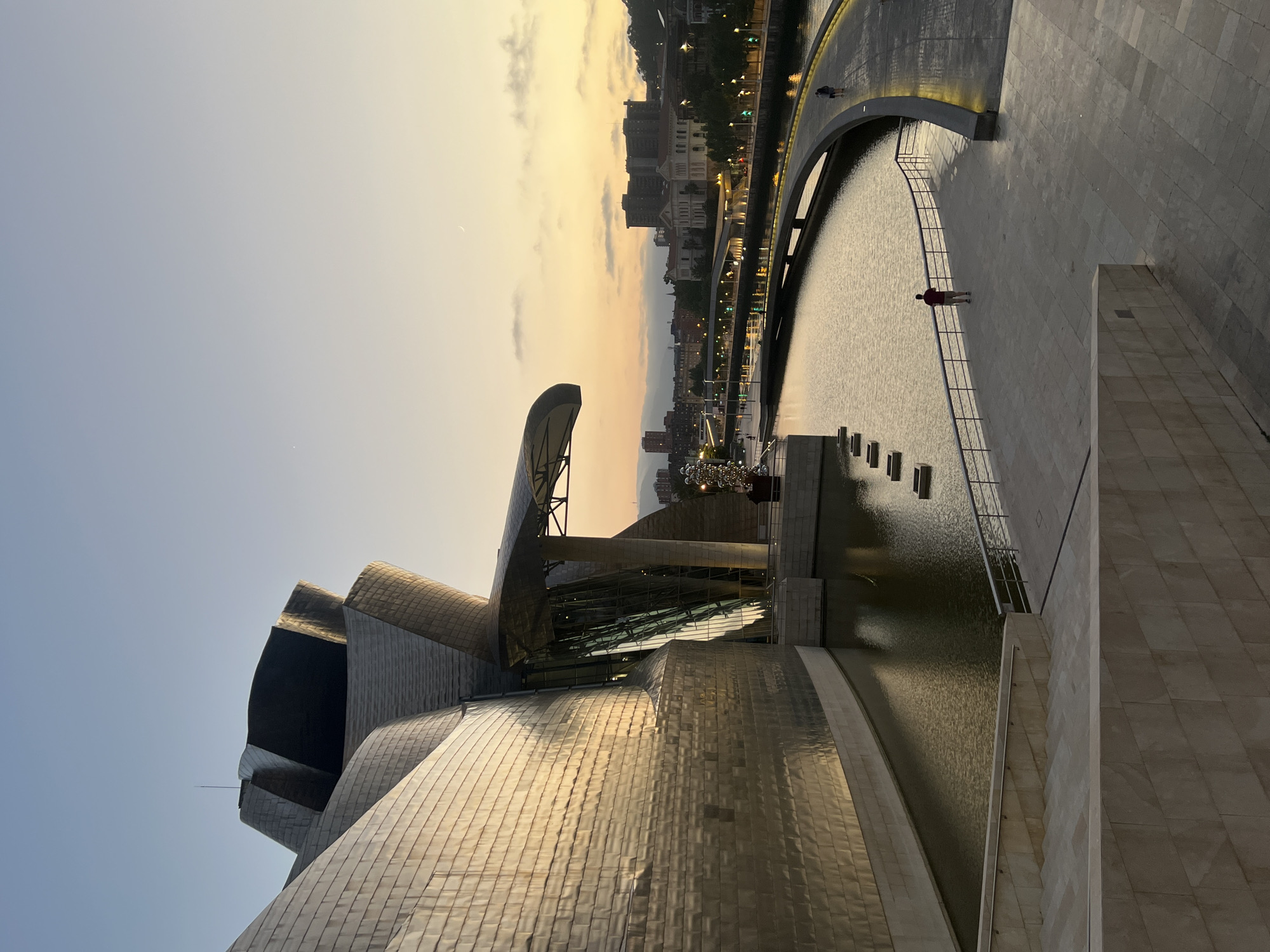 The image captures the iconic Guggenheim Museum Bilbao at dusk, a globally recognized architectural landmark in Bilbao, Spain. The museum's exterior, clad in shimmering titanium, reflects the soft, warm hues of the setting sun, creating a dramatic visual effect. Its distinctive, fluid architecture, characterized by curved surfaces and interlocking volumes, dominates the left side of the frame.
In the foreground, light-colored stone steps and a wide promenade lead down to the museum's reflective pool. A single person, dressed in a red shirt and dark shorts, stands on the promenade, looking out towards the river. The Nervión River flows beside the museum, its surface calm and mirroring the twilight sky and the city lights that are beginning to illuminate the opposite bank.
A gracefully curving pedestrian bridge, edged with lights, spans the river, connecting the museum area to the cityscape beyond. Several figures are visible on the bridge, including two individuals walking together. On the far bank, the city of Bilbao unfolds with a mix of architectural styles, including classical buildings and more modern structures, all silhouetted against the fading light. The sky above is a gradient of soft blues and yellows, with a faint crescent moon visible. The overall atmosphere is serene and majestic, highlighting the interplay of light, architecture, and the urban environment.