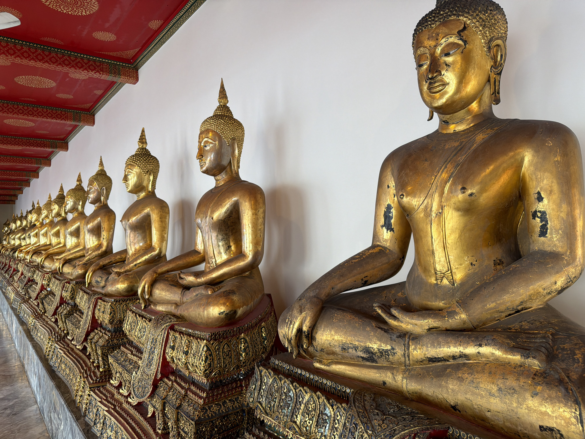 In a long, covered walkway within a Buddhist temple in Bangkok, Thailand, a row of at least ten gilded Buddha statues are seated in meditation. The statues are arranged in a line and are viewed from a slightly elevated, close-up perspective, with the closest statue occupying the right foreground. The statues are made of a material that appears to be gilded metal, showing some wear and discoloration. They are adorned with traditional robes and sit on ornate, multi-tiered bases decorated with intricate gold carvings and red fabric. The background consists of a plain white wall and the underside of a red ceiling adorned with golden patterns. The floor is a polished marble or stone. The lighting suggests an indoor setting with ambient light. The overall impression is one of reverence and historical artistry.