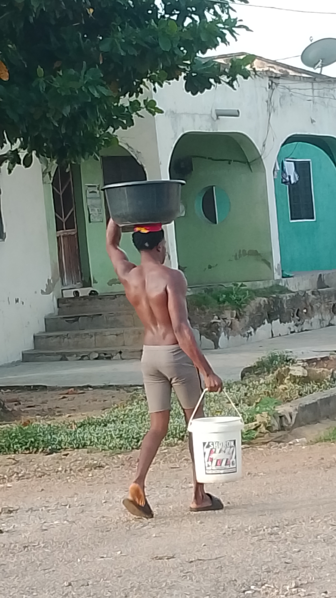 The image captures a young, muscular man walking away from the camera on a dirt or gravel path, suggesting a rural or semi-urban setting, consistent with Lokoja, Nigeria. He is shirtless, displaying a well-defined back, and wears light-colored shorts and dark flip-flops.
The man is engaged in transporting items, balancing a large dark grey or black basin on his head. A colorful piece of fabric, primarily red and yellow, is visible between his head and the basin, likely providing cushioning. In his right hand, he carries a white plastic bucket with a handle. On the side of the bucket, the text "SHOT ON" is clearly visible, followed by "ITEL" and additional, indistinct lettering or a logo, suggesting it's a watermark from a mobile phone camera.
The background features residential buildings under a bright sky, indicating daylight. One building on the left has light-colored walls with green accents around arched doorways and windows, and a set of concrete steps leading up to a door. Another structure to the right is painted a distinct teal-green color, with a circular cut-out detail and a window where some laundry appears to be hanging. Satellite dishes are visible on the roofline of one of the buildings. Green foliage from trees partially obscures the upper left portion of the scene. The overall impression is one of daily life and physical labor in a warm, sunny environment.