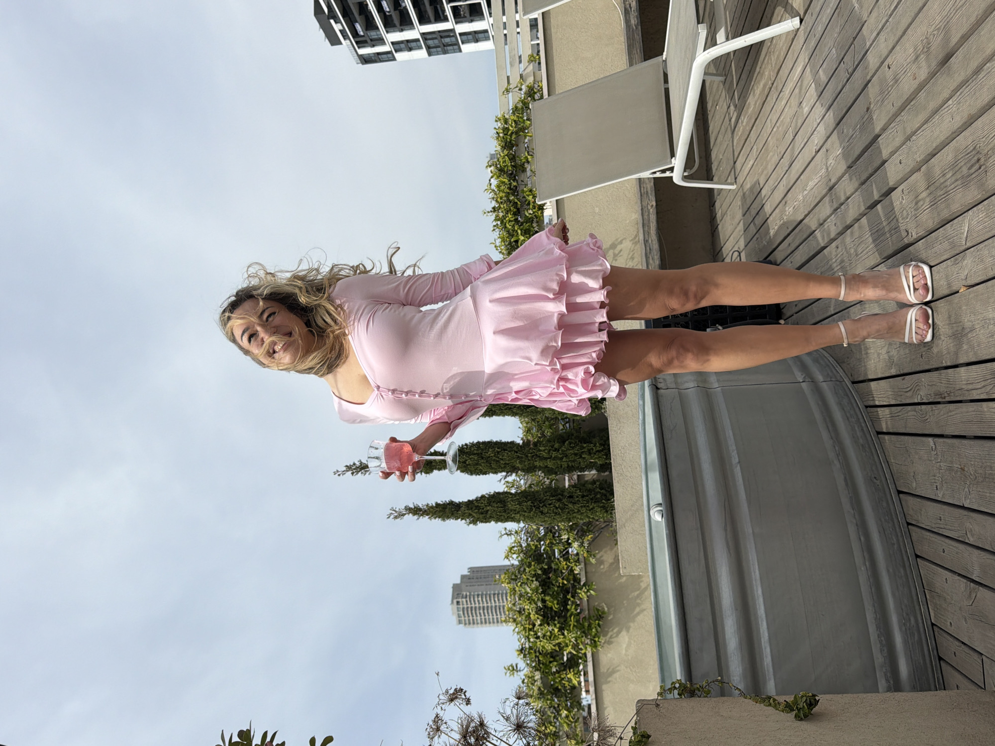 A young woman stands on a wooden deck, holding a glass of pink liquid, possibly rosé. She is wearing a light pink, long-sleeved mini dress with a ruffled skirt, and white strappy heeled sandals. Her blonde hair is loose and wavy. Behind her are several tall, thin evergreen trees, a grey building, and another building in the distance. The sky is overcast. Next to her on the deck are a couple of white metal chairs and a grey tub. Based on the presence of buildings with specific architectural elements and the vegetation style, the location can be approximated in Tel-Aviv, Israel.