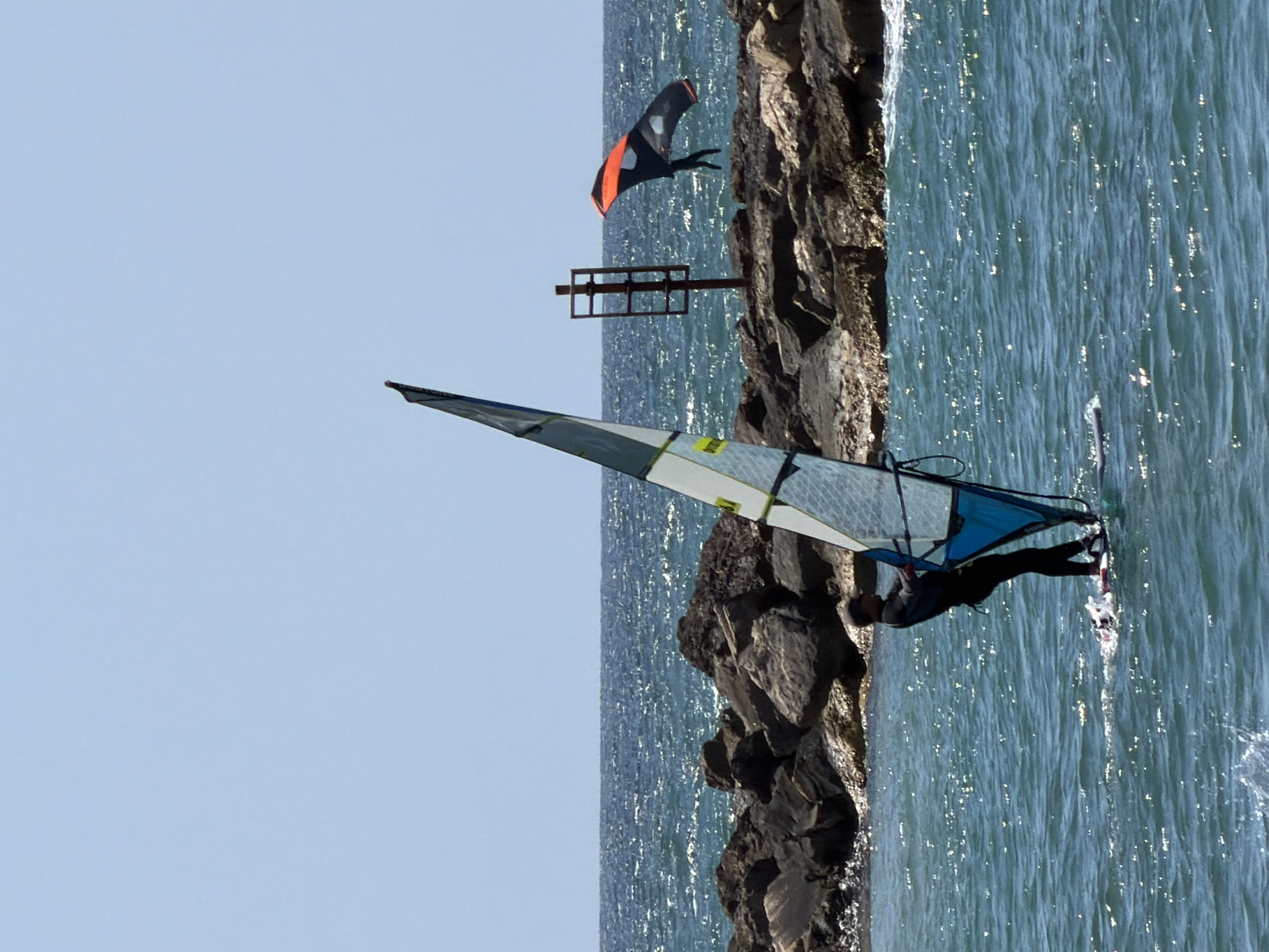 The image shows a watersport activity near a rocky shore in Tel-Aviv, Israel. A windsurfer is on the water, with the sail positioned at an angle. Another person with a kite is visible in the background, closer to the rocks. The water reflects sunlight. A metal structure is also visible in the background, standing on the rocks. The sky is clear and blue.