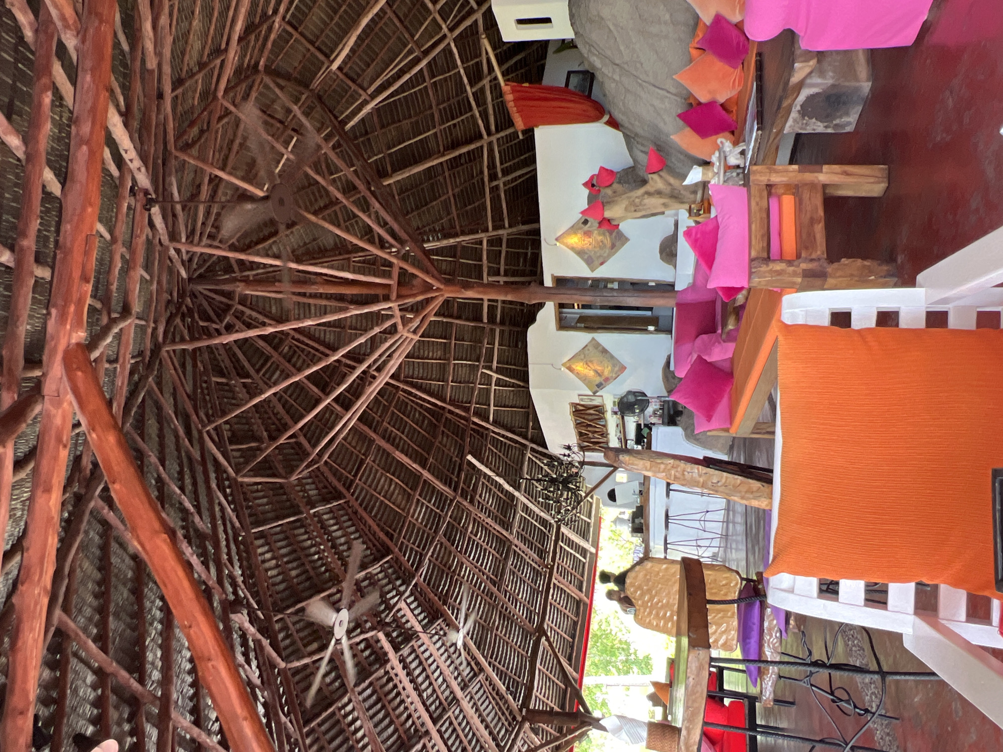The image shows an indoor seating area with a thatched roof supported by wooden beams. Two ceiling fans are visible near the roof. The area is decorated with bright pink and orange cushions on various wooden furniture. In the background, there is a white counter with stools and a few people are standing near it. The floor is a deep red color. There are some large rocks incorporated into the seating area design. The overall impression is of an open-air lounge or restaurant. The location is identified as Baan Chaweng Noi, Thailand.