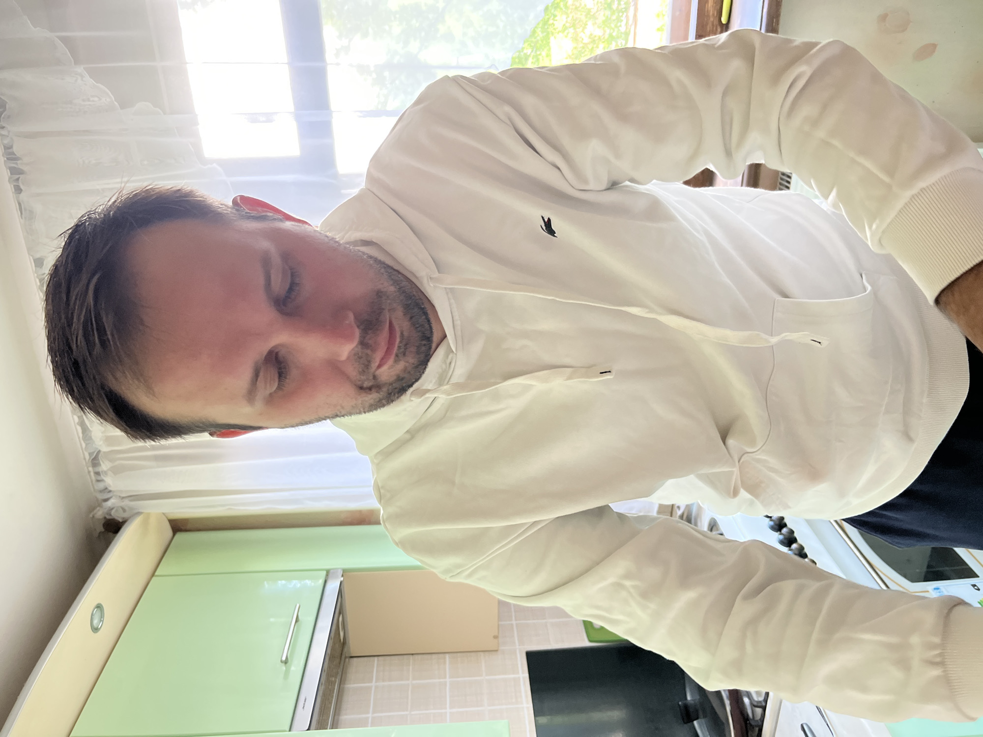 A man with short brown hair and a beard, wearing a white hooded sweatshirt with a small, dark logo on the chest, is looking down. He is standing in a kitchen with light green cabinets, a beige tile backsplash, and a window in the background with bright light shining through. A black object is visible on the countertop. The location is likely a residence in Rusanovo, Russia, given the prompt.