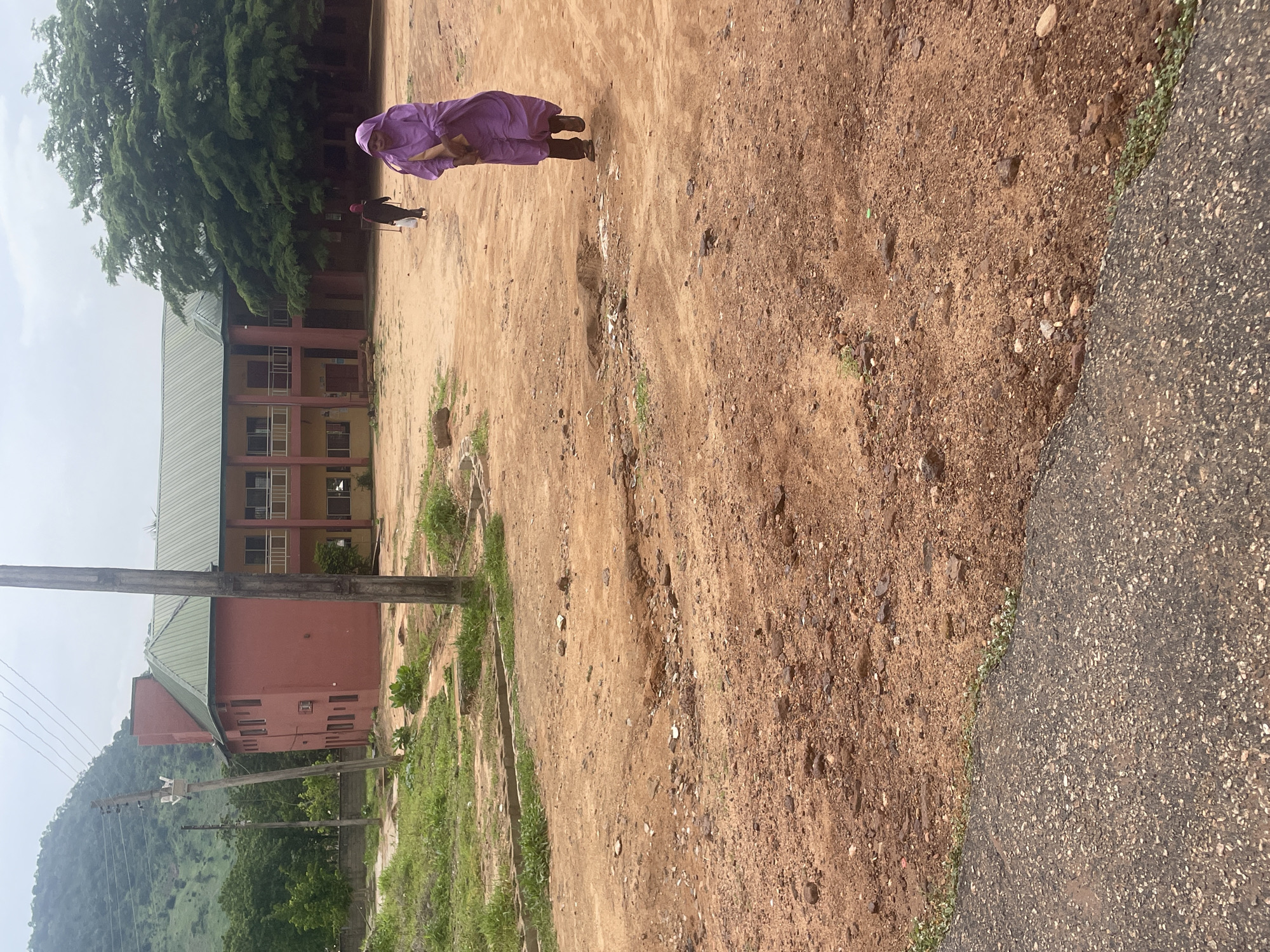The image depicts an outdoor scene on a cloudy day, likely in Lokoja, Nigeria, judging by the building style and vegetation. In the foreground, a person dressed in a bright purple abaya is walking away from the camera across a dusty, uneven ground. Behind them, another person in dark clothing is also walking away. To the left, a large, two-story building with a green roof and red and yellow walls is visible, with a hillside covered in vegetation and trees in the background. Several utility poles with wires are present throughout the scene, one standing prominently in the center. The ground is a mixture of dirt, small stones, and sparse patches of grass. The overall environment suggests a somewhat rural or semi-urban setting.