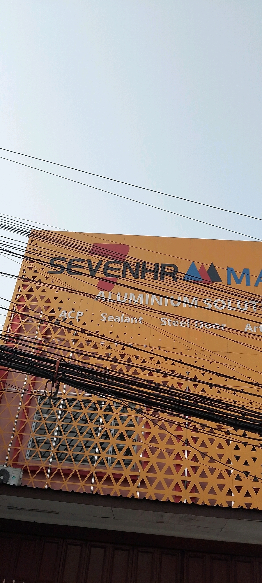 In Bekasi, Indonesia, the image captures the facade of a building with an orange exterior. The building features a prominent sign with the text "SEVENHR ALUMINIUM SOLUT". Below this, smaller text reads "ACP Sealant Steel Door Art". The facade is decorated with a geometric pattern of triangles. Numerous black electrical wires are strung across the front of the building, obscuring parts of the signage and decorative elements. A window with a similar triangular pattern behind it is visible on the left side of the building. An air conditioning unit is mounted to the wall near the window. The sky above is overcast.