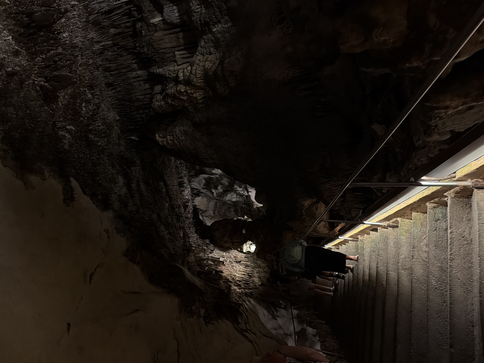 The image captures a dimly lit interior, likely a cave, in the city of Manacor, Spain. The foreground features rough, textured walls and stalactite formations, typical of cave environments. A set of stone steps, partially illuminated by artificial lighting, descends into the depths of the cave. A metal railing runs parallel to the stairs, providing support. Several people can be seen walking down the stairs, their forms silhouetted against the ambient light. The overall environment is dark and rugged, with a focus on the natural geological formations of the cave.