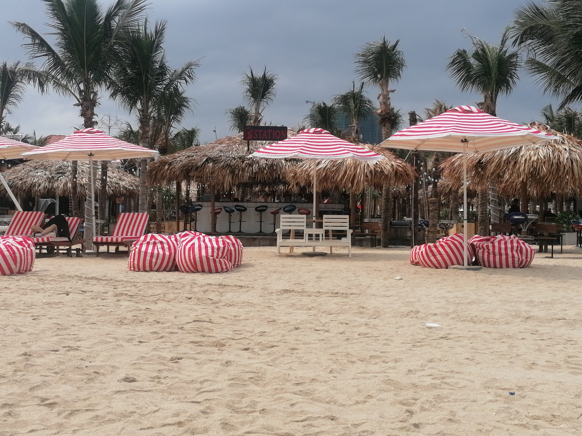 The image displays a relaxed beach club scene on a sandy shore, likely in Bai Chay Ward, Vietnam, under an overcast sky. The foreground is dominated by light brown sand, showing subtle textures and footprints.
Several red and white striped bean bags are scattered on the sand, offering informal seating. In the midground, multiple large beach umbrellas with matching red and white stripes provide shade. Beneath one umbrella to the left, a person is visible relaxing on a striped beach lounger, with only their lower body and crossed legs, clad in dark clothing, clearly discernible. Further into the midground, there are more striped loungers.
A central element is a thatched-roof structure, serving as a bar or service point. A red sign mounted on this structure prominently displays the word "STATION". In front of the station, several dark bar stools are arranged. To the right of the station, a small white wooden table is paired with two white chairs, forming another seating area. More thatched structures are partially visible behind and beside the main station.
The background is characterized by numerous tall palm trees with green fronds, indicative of a tropical setting. The sky is a uniform light grey, suggesting an overcast or cloudy day. The overall scene conveys a tranquil, leisurely atmosphere typical of a beach resort or club. No specific event is actively taking place, implying a quiet period or anticipation of guests.