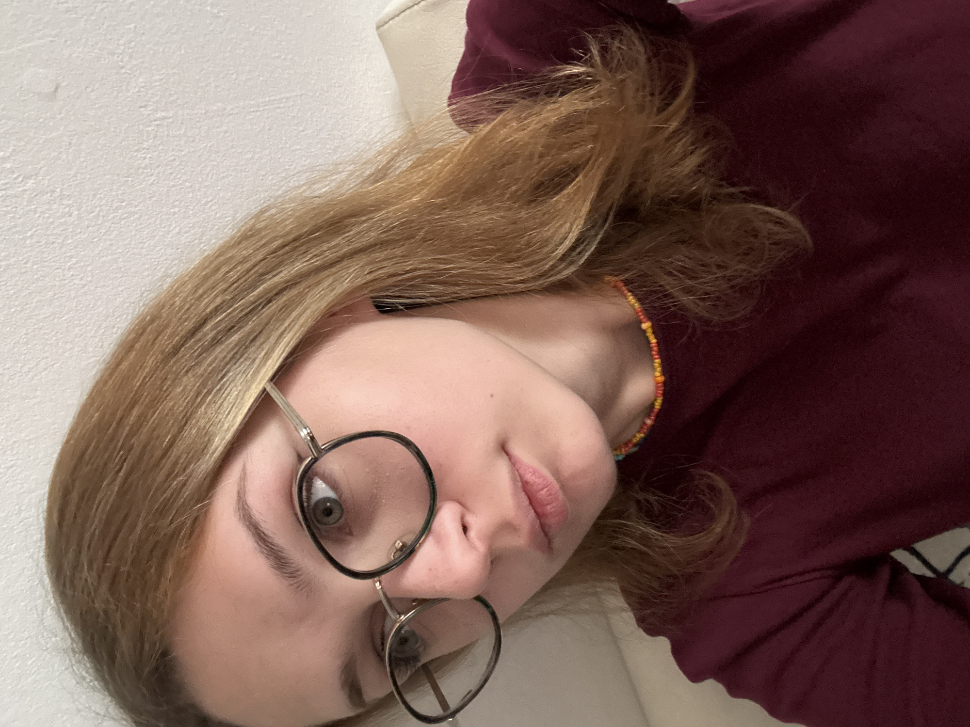 A close-up, slightly rotated photograph shows a young woman with blonde hair, wearing round, wire-framed glasses. She is wearing a maroon-colored shirt and a colorful beaded necklace. The background appears to be a white wall and a white pillow. It is not possible to determine the location as Kragujevac, Serbia from the image provided.