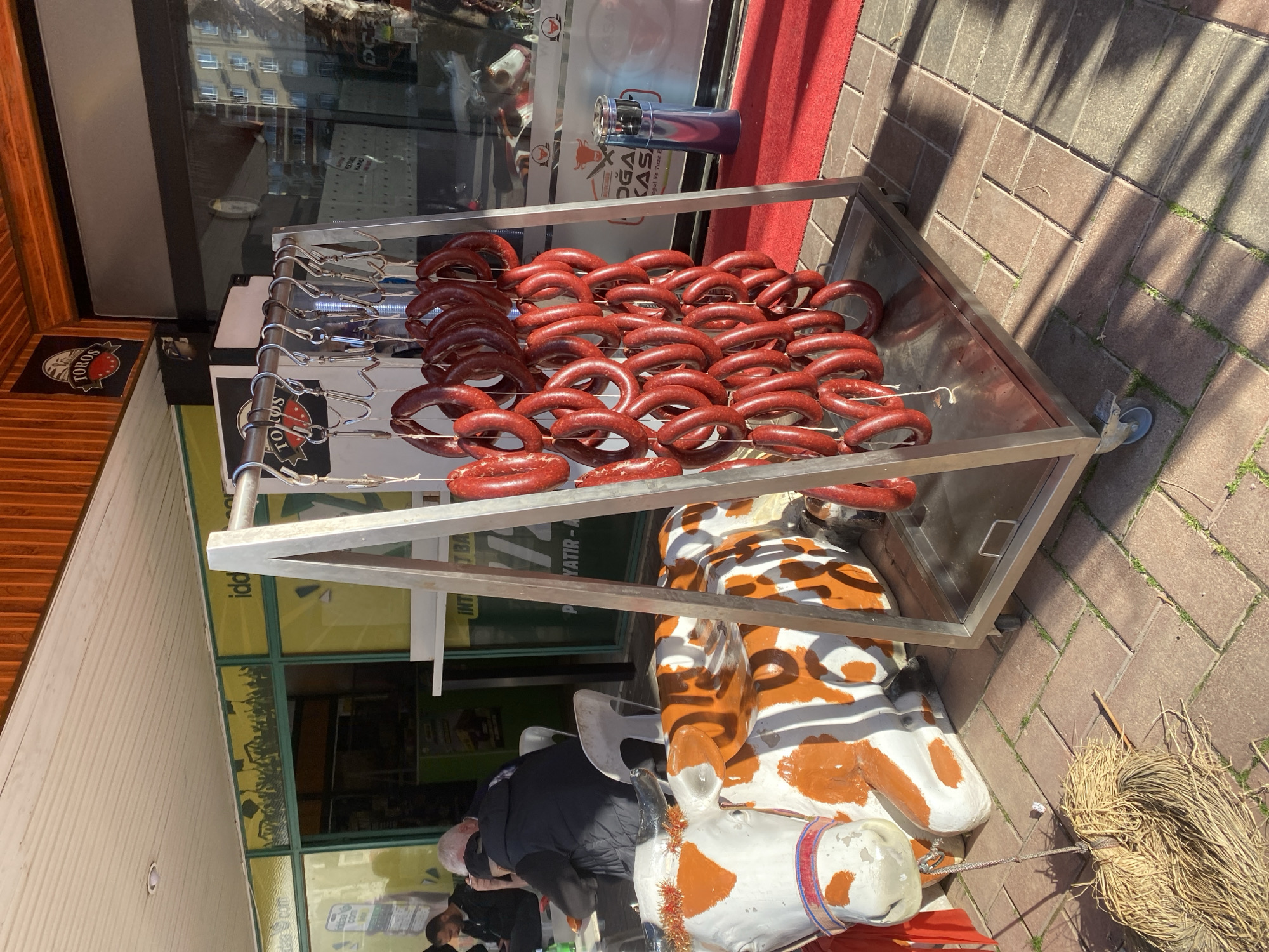 The image captures a street scene in Alanya, Turkey, displaying a metal cart filled with numerous red sucuk sausages hanging on hooks. The cart is positioned outside a shop, and a decorative cow statue lies next to it. Individuals are present in the background. The surroundings include brick pavement, a red carpet, and visible shop windows.