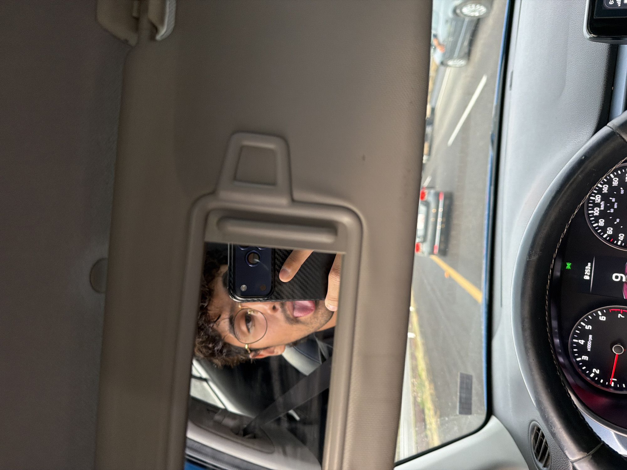 A selfie reflection in a car visor mirror. The man is holding a smartphone, sticking out his tongue. The reflection includes a partial view of the driver's side window, the steering wheel, and the dashboard. Outside the car, a street with other vehicles and lane markings can be seen. The car's speedometer reads 9 km/h, and the odometer is 253 km. The specific location within San Bernardino Chalchihuapan, Mexico is not identifiable from the image.