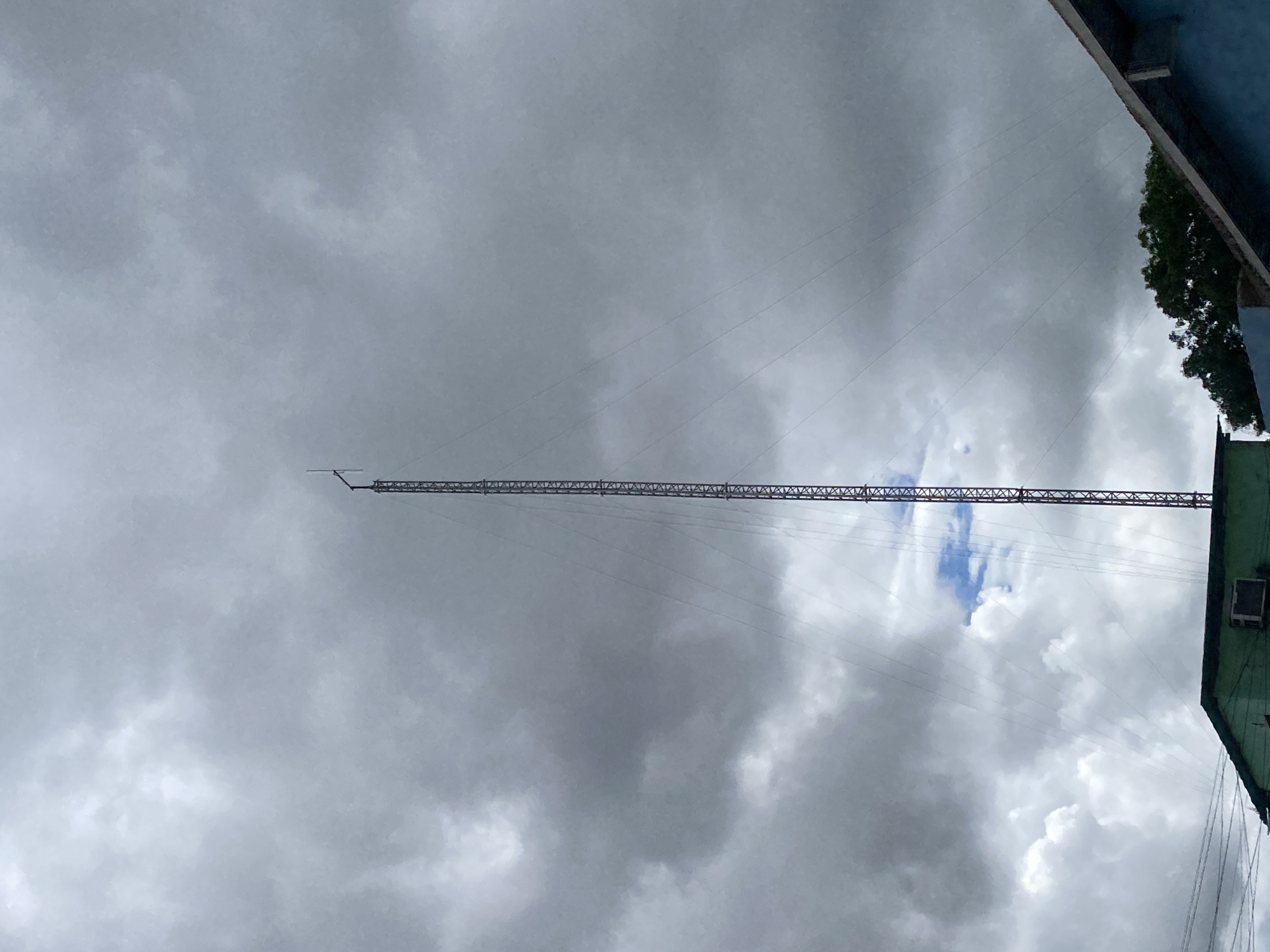 The image depicts a low-angle view in Tinaquillo, Venezuela, showing a tall, slender metal communications tower extending upwards into a cloudy sky. The tower is held in place by guy wires. Fragments of buildings, including a rooftop and trees, are visible in the upper right corner of the image. The sky is predominantly gray with various cloud formations and a small patch of blue visible behind the tower.