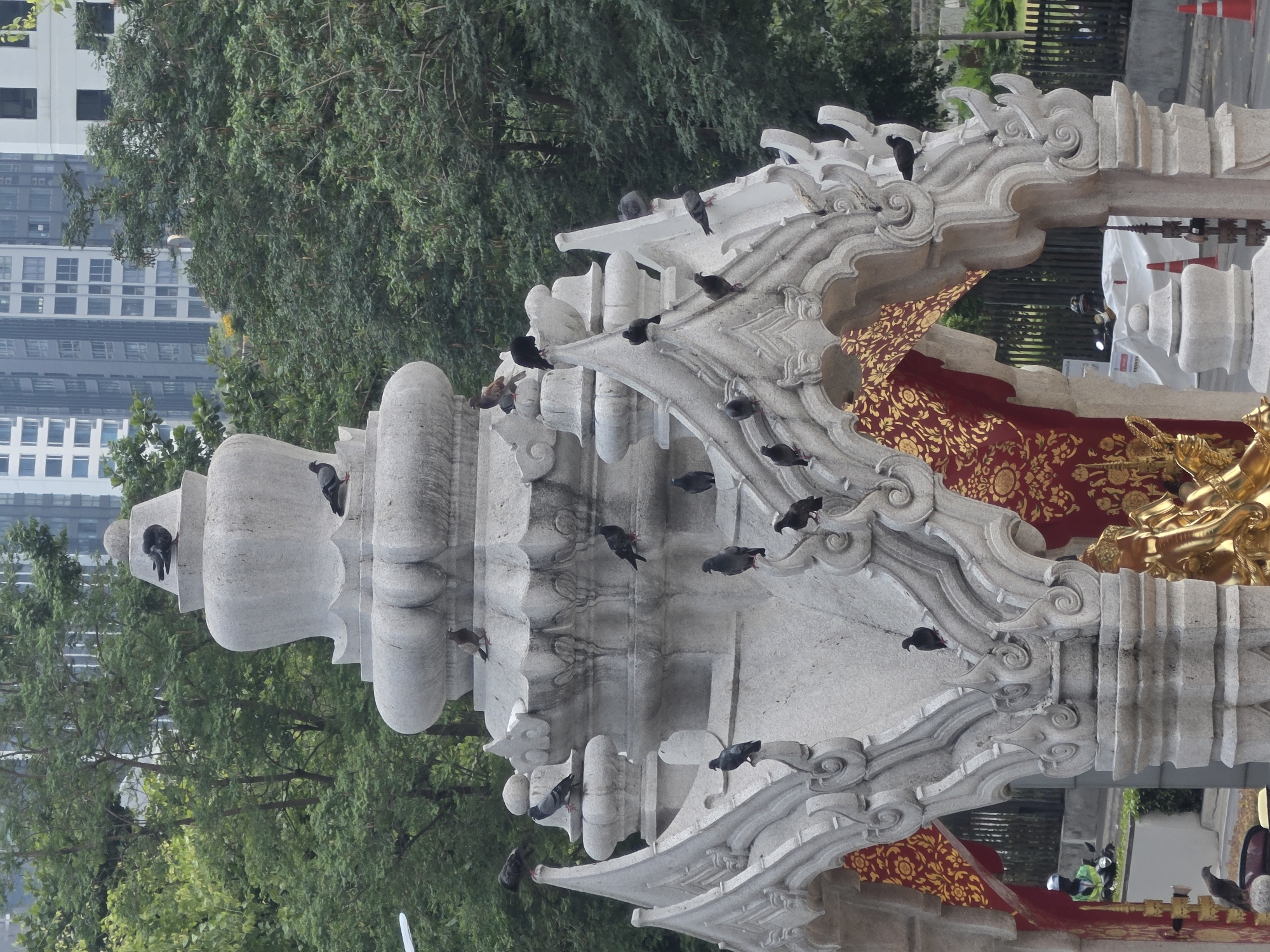 The photograph depicts an ornately carved, light-colored stone or concrete structure, likely part of a temple or religious building, situated in an urban environment. Pigeons are perched on various parts of the structure. Red and gold decorative fabric is draped within the structure.
In the background are green trees and a tall, modern building with numerous windows, indicative of an urban setting. The presence of the structure, the surrounding environment, and the architectural style are characteristic of Bangkok, Thailand.