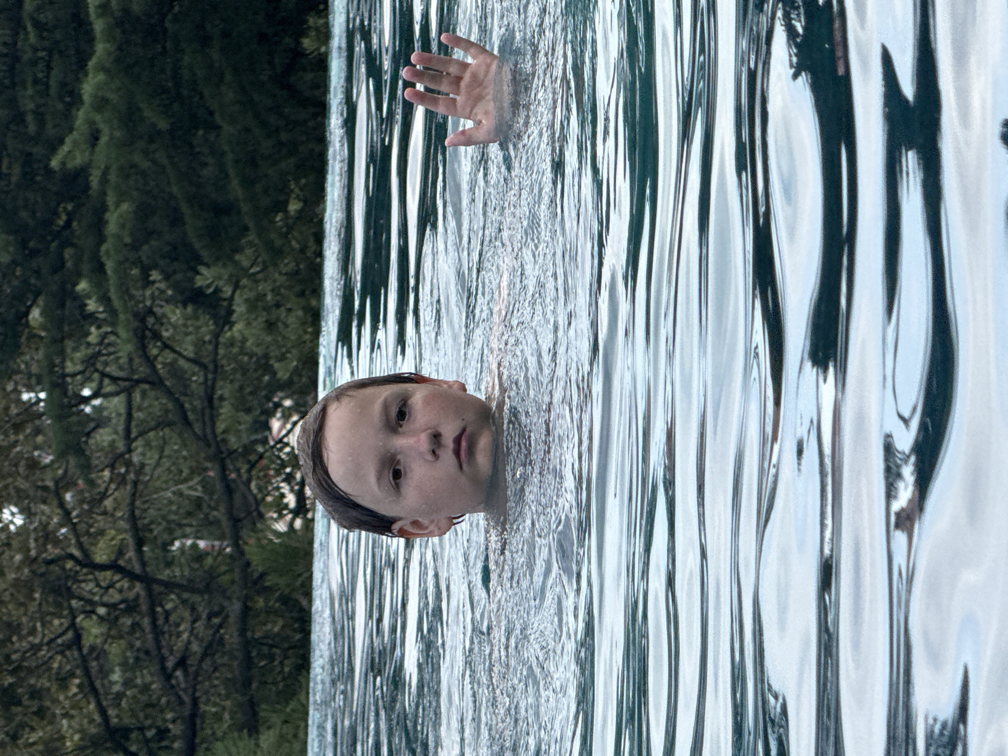 A young boy with wet hair is in a swimming pool. He is looking directly at the camera with a serious expression. His right hand is raised out of the water, palm facing forward. The pool water is blue and has ripples. In the background, there are trees. The setting does not provide any specific clues to indicate it is located in the city of Yalta, Ukraine.