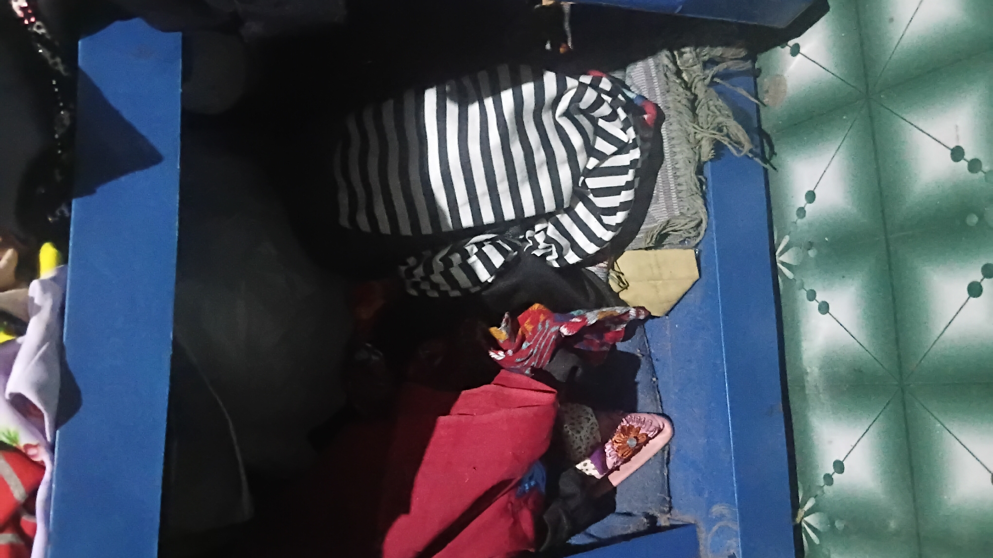 The image shows a cluttered shelf inside what appears to be a residential storage area. The shelf is painted blue and is filled with various items of clothing and fabric. On the left side of the shelf, a red garment is prominent, along with a pink slipper decorated with flowers. To the right of the red garment, a black and white striped fabric item is folded. Beneath these items, a gray fringed textile is visible. The background of the shelf appears dark and indistinct. The floor beneath the shelf is tiled with a green and white geometric pattern. The scene is dimly lit, suggesting it may be indoors, possibly a bedroom or a storage room. The context provided by the prompt places this scene in Prabumulih, Indonesia.
