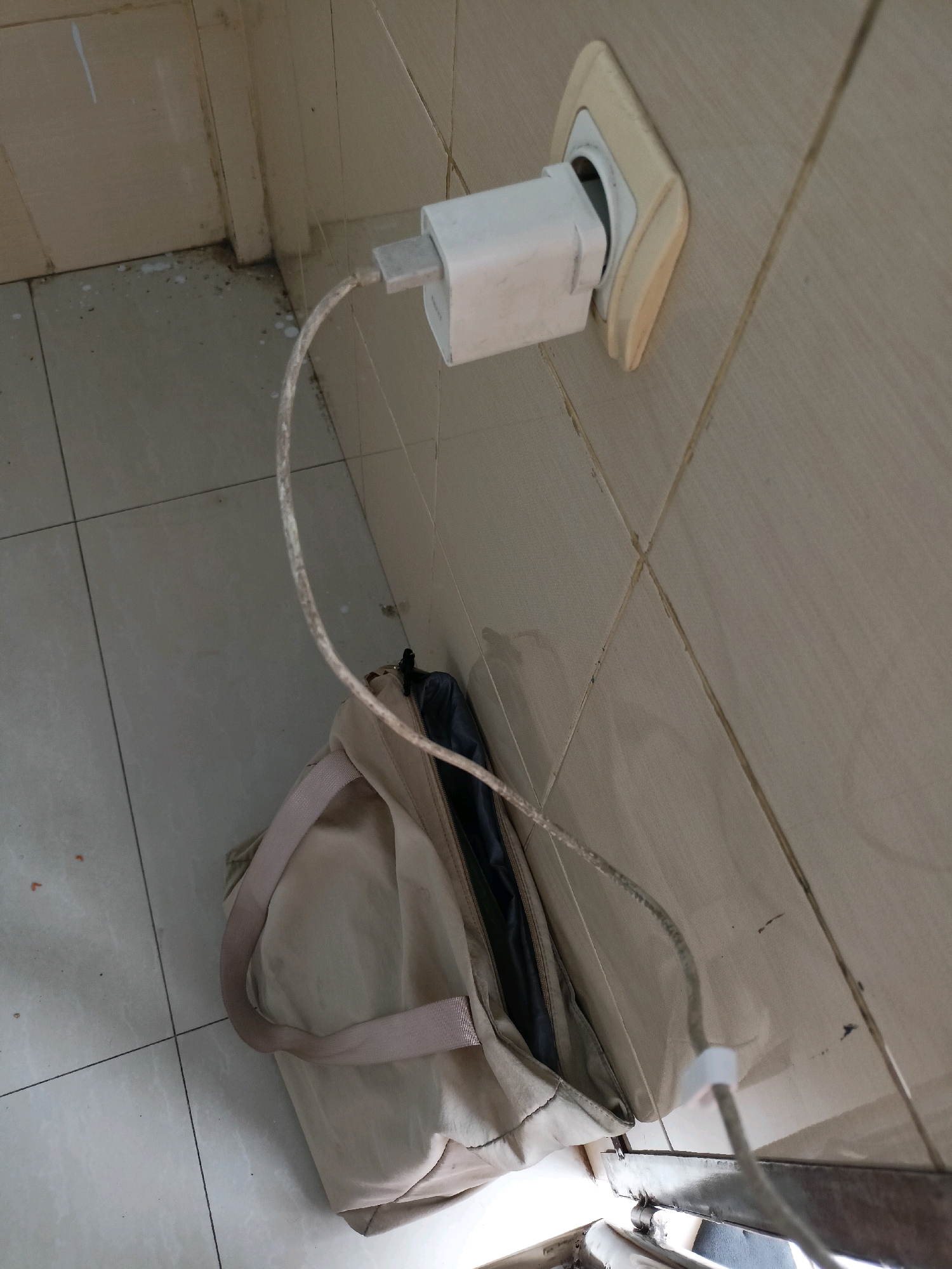 A white charging brick is plugged into a cream-colored wall outlet. A gray charging cable extends from the brick and drapes over a beige duffel bag. The bag is open and appears to be made of nylon. The scene is indoors, on a tiled floor and wall, possibly in Wancen, Indonesia, given the outlet type. The lighting is dim.