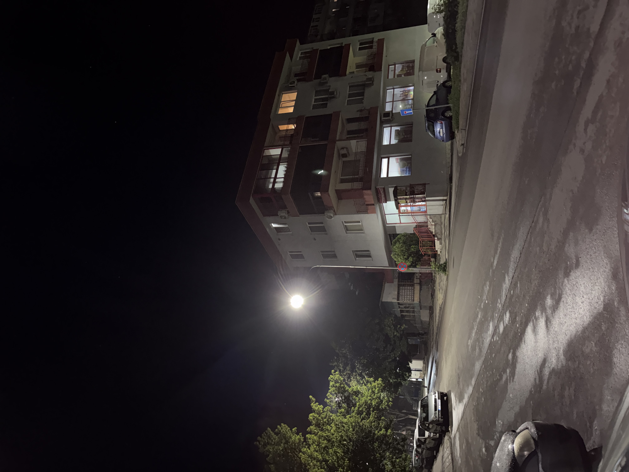 The image captures a quiet urban street scene at night, likely in Shumen, Bulgaria, based on visible text. The photograph is oriented vertically, showing a multi-story building dominating the right side of the frame. This building features a light-colored facade with numerous windows, balconies accented in a reddish-brown hue, and visible air conditioning units. Several windows are illuminated from within, suggesting occupied apartments or offices.
A bright streetlamp in the mid-left casts a strong, starburst-like glow across the scene, illuminating parts of the building, the street, and nearby trees. The street itself, positioned to the left and extending into the foreground, appears wet or reflective, possibly from recent rain, showing patches of light and dark. Several cars are parked along the street; a blue car is clearly visible near the building, and other vehicles are discernable further down. Lush green trees are visible beneath and around the streetlamp, adding organic elements to the urban setting. The sky is dark and clear, with a few faint specks of light that could be stars or distant light sources.
Notable details include:
- A red and white circular prohibitory road sign (likely "No Stopping" or "No Parking") attached to a pole near the building's entrance.
- A blue rectangular road sign (possibly a direction or information sign) further up the building facade.
- A sign on the ground floor of the building clearly displays "СУПЕРМАРКЕТ" (SUPERMARKET) in Cyrillic, providing a strong geographical cue.
The scene evokes a sense of calm and stillness typical of an urban environment late at night, with artificial lights defining the visible space. No people or specific activities are observed.