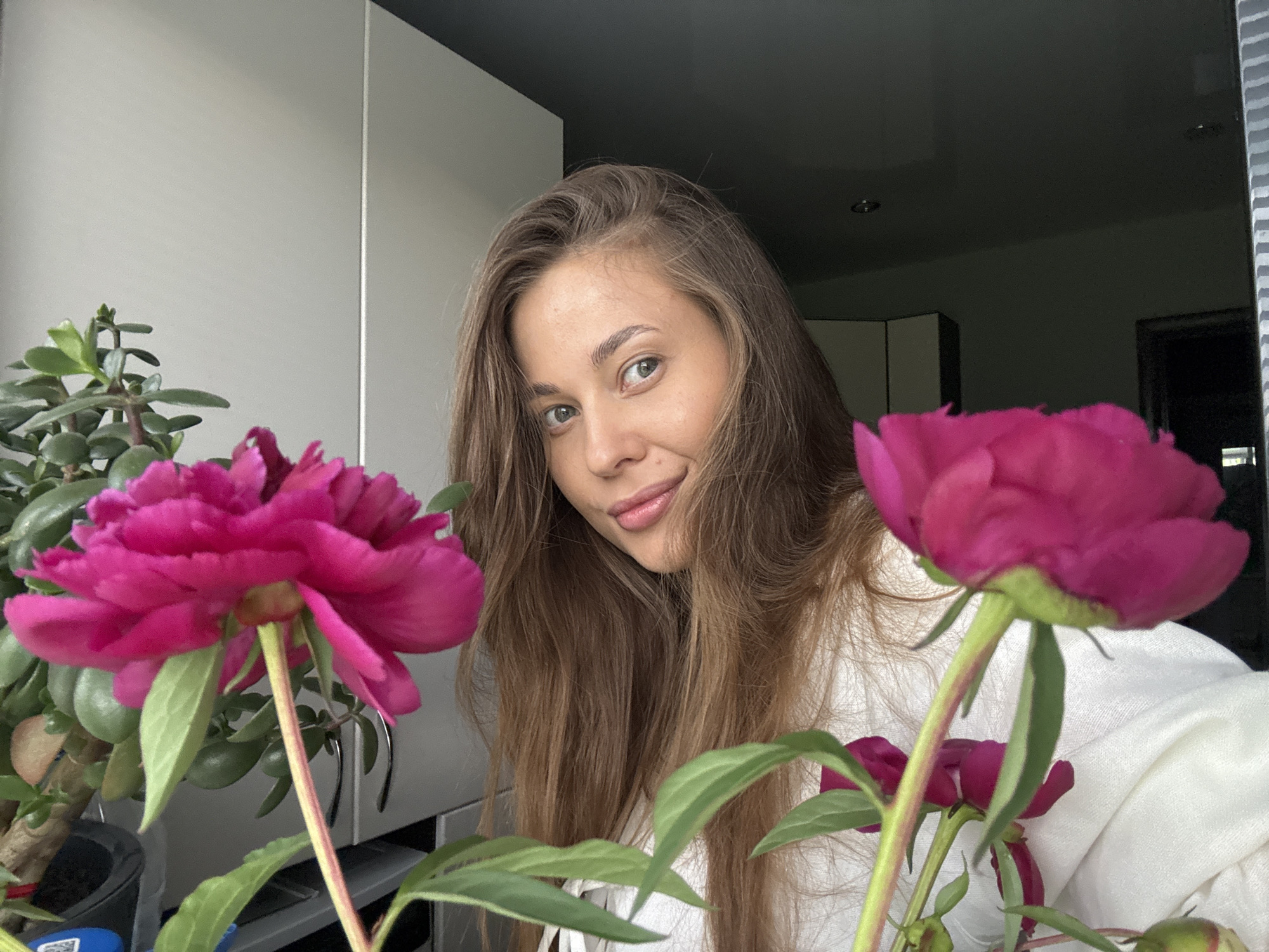A young woman with long brown hair, wearing a white shirt, is framed by bright pink peonies and a jade plant. She is positioned in the center of the frame, looking directly at the camera with a slight smile. The background appears to be an indoor setting with light-colored cabinets and a dark ceiling. The image's lighting suggests it was taken during the daytime, possibly near a window. The overall composition is intimate and natural, with the flowers adding a touch of color and softness. There is no information in the image to determine the location in the city of Chernushka, Russia.