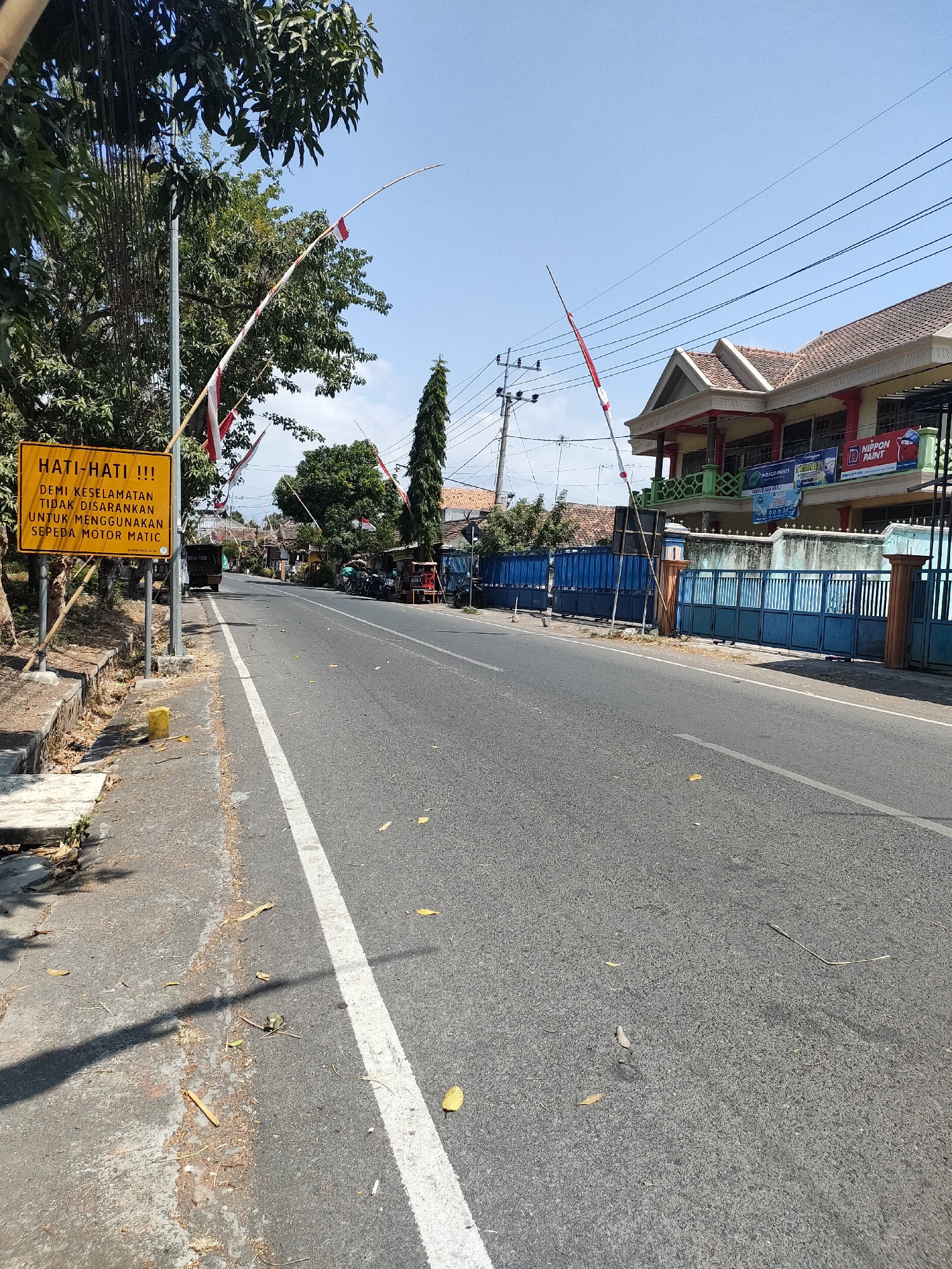 The image captures a street scene in Tongaswetan, Indonesia, on a clear day. A yellow warning sign is prominently displayed on the left side of the road, with the text "HATI-HATI !!! DEMI KESELAMATAN TIDAK DISARANKAN UNTUK MENGGUNAKAN SEPEDA MOTOR MATIC." This translates to "BE CAREFUL !!! FOR SAFETY, IT IS NOT RECOMMENDED TO USE AUTOMATIC MOTORCYCLES." The sign is supported by metal posts and is accompanied by Indonesian flags.
The street itself is paved and appears to be a two-lane road. On the right side of the street, a building with a red and yellow facade and a tiled roof stands, featuring signage for "NIPPON PAINT" and "INDIGO PAINTS." A blue metal gate encloses the property in front of the building. Various trees, including a tall, slender evergreen, are visible along the street. Several motorcycles are parked on the side of the road near the building. A truck is also seen driving down the road in the distance. The ground is littered with fallen leaves and debris, suggesting a natural environment adjacent to the urban setting. Overhead, power lines crisscross the sky. The overall impression is a typical roadside view in a residential or commercial area of an Indonesian town.