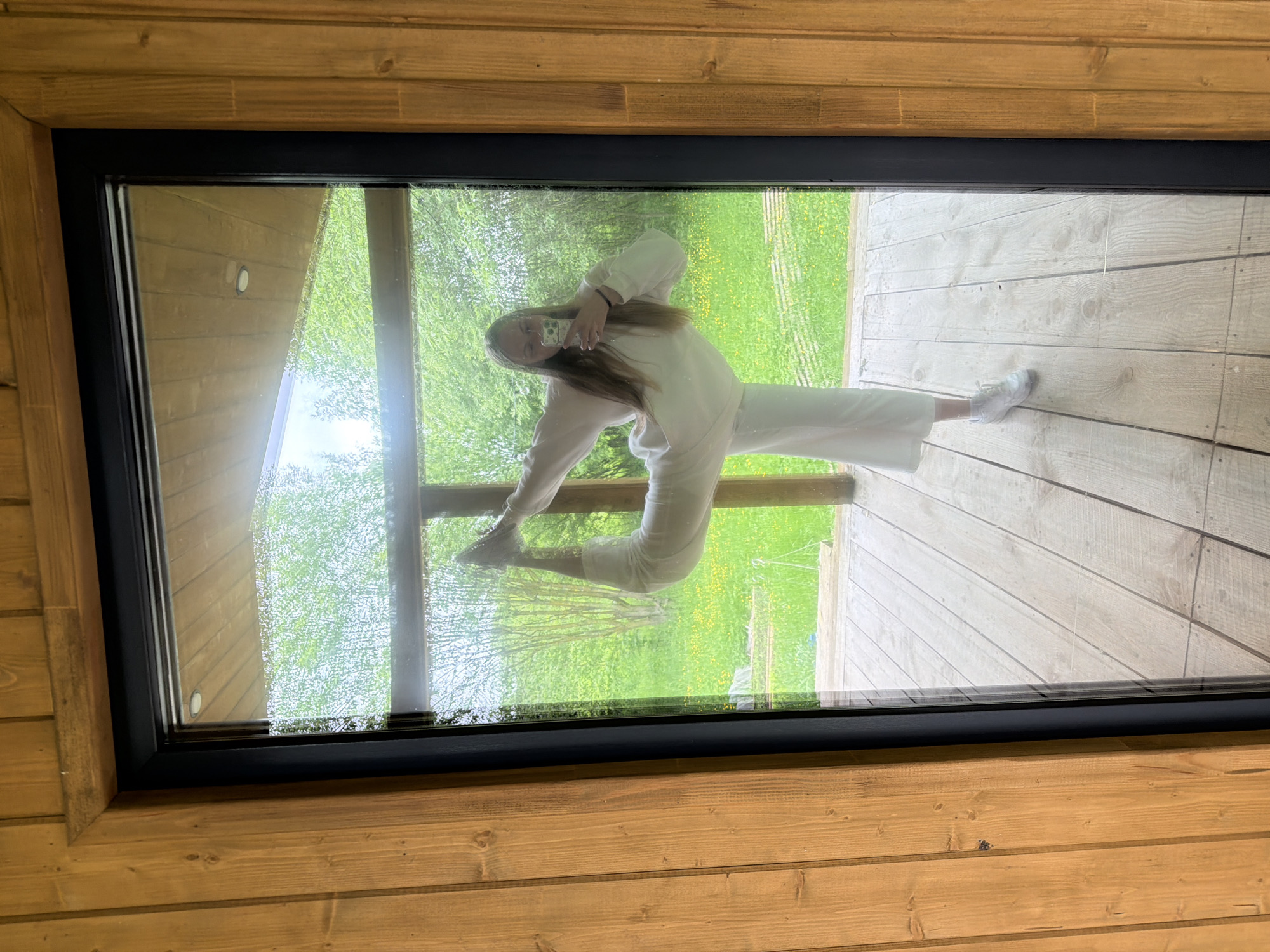 A young woman is captured in a dynamic pose, seemingly performing a split, as she takes a selfie reflected in a large, rectangular glass panel. The image is taken from above, looking down onto the window, which is framed by a black border and set into a light-colored wooden structure, possibly a floor or ceiling skylight in a building in Vozhoiskii, Russia. The wooden planks feature a natural grain.
The woman has long, light brown hair and is dressed in a light-colored, casual tracksuit or loungewear. She holds a silver smartphone in her right hand, pointed towards the glass to capture her reflection. Her pose is unusual, with one leg extended upwards in a split-like fashion and the other bent, creating an illusion that she is suspended or balancing. Her reflection appears to merge with the outdoor scenery.
Through the clear glass, a vibrant green natural environment is visible, featuring lush trees, grass, and scattered yellow flowers, indicative of a bright, daytime setting. A light-colored wooden deck or pathway can be seen immediately outside the window, where the woman's reflected leg seems to touch. The scene is well-lit, suggesting clear weather. No visible text is present in the image.