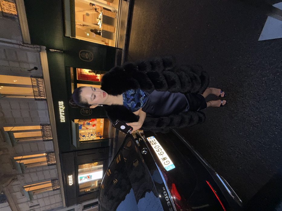 A stylish woman stands gracefully on a street at night, positioned next to a sleek black car. She is the central subject, wearing a voluminous black furry coat over a shimmering dark blue top and a dark skirt, complemented by open-toed sandals. Her dark hair is neatly styled in a bun, and she wears delicate earrings. She holds a smartphone in her left hand, which displays an active call interface.
Beside her, a glossy black Mercedes-Benz E 300e is parked, identifiable by the "E 300e" emblem on its trunk and the French license plate "GW-650-RE." The car's polished surface reflects the ambient lights.
In the background, an elegant commercial building, likely a luxury boutique, is brightly lit. The prominent signage "DIPTYQUE" is clearly visible on its facade, and a fainter "CELINE" sign can be discerned in a lower window. The ground appears wet, suggesting recent rain. The setting evokes a sophisticated urban evening ambiance, characteristic of Paris, France, given the visible brand names and license plate.