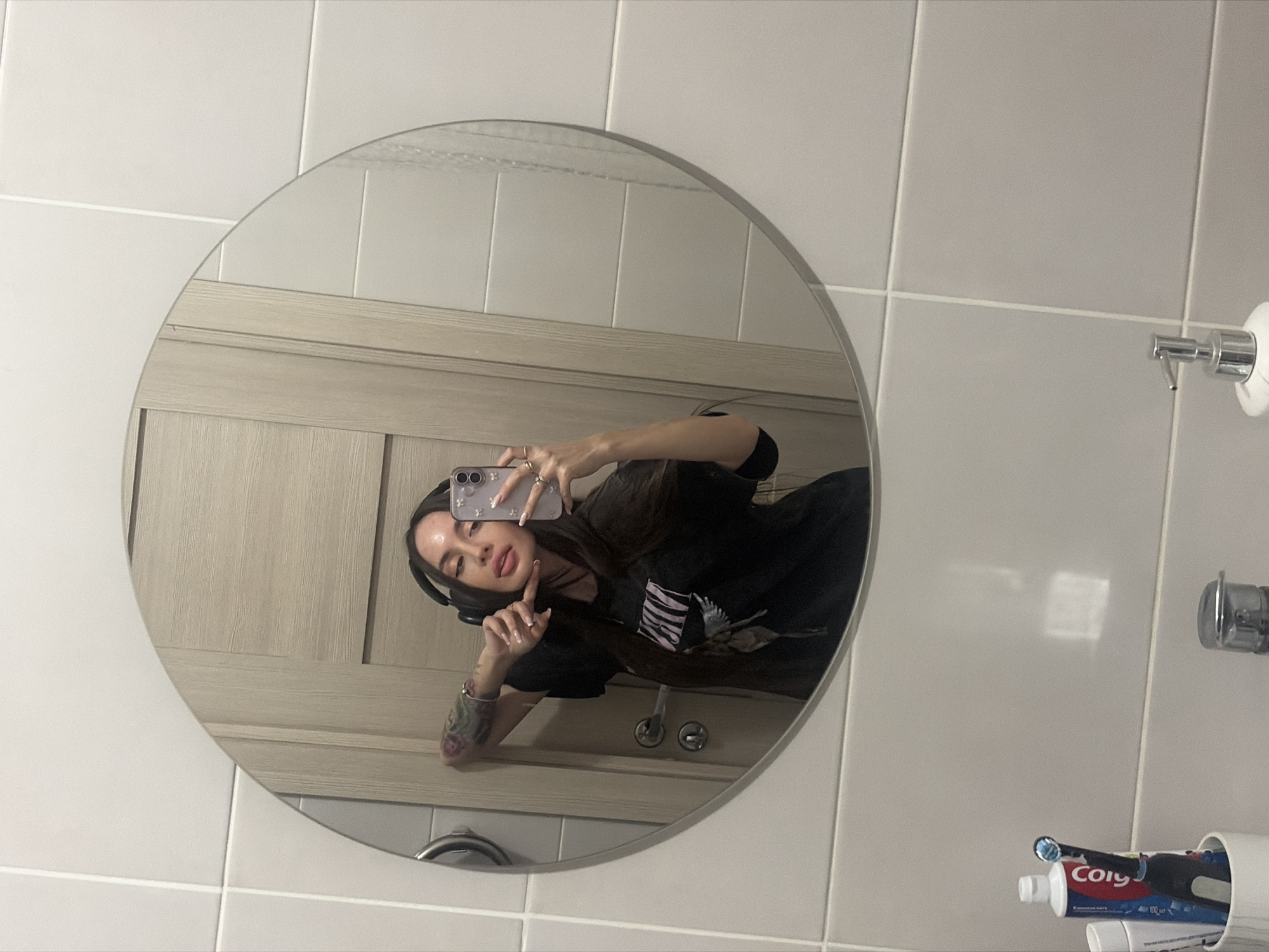 A selfie reflection of a woman in a round mirror. The woman has long brown hair, headphones, and a black shirt with a bird design. She is holding a phone with a floral case and several rings on her fingers. The background shows a light-colored wooden door, white tiled walls, a soap dispenser, and a toothbrush holder with Colgate toothpaste. The location is likely a bathroom, but no specific location in Saint Petersburg, Russia, can be determined from the image.