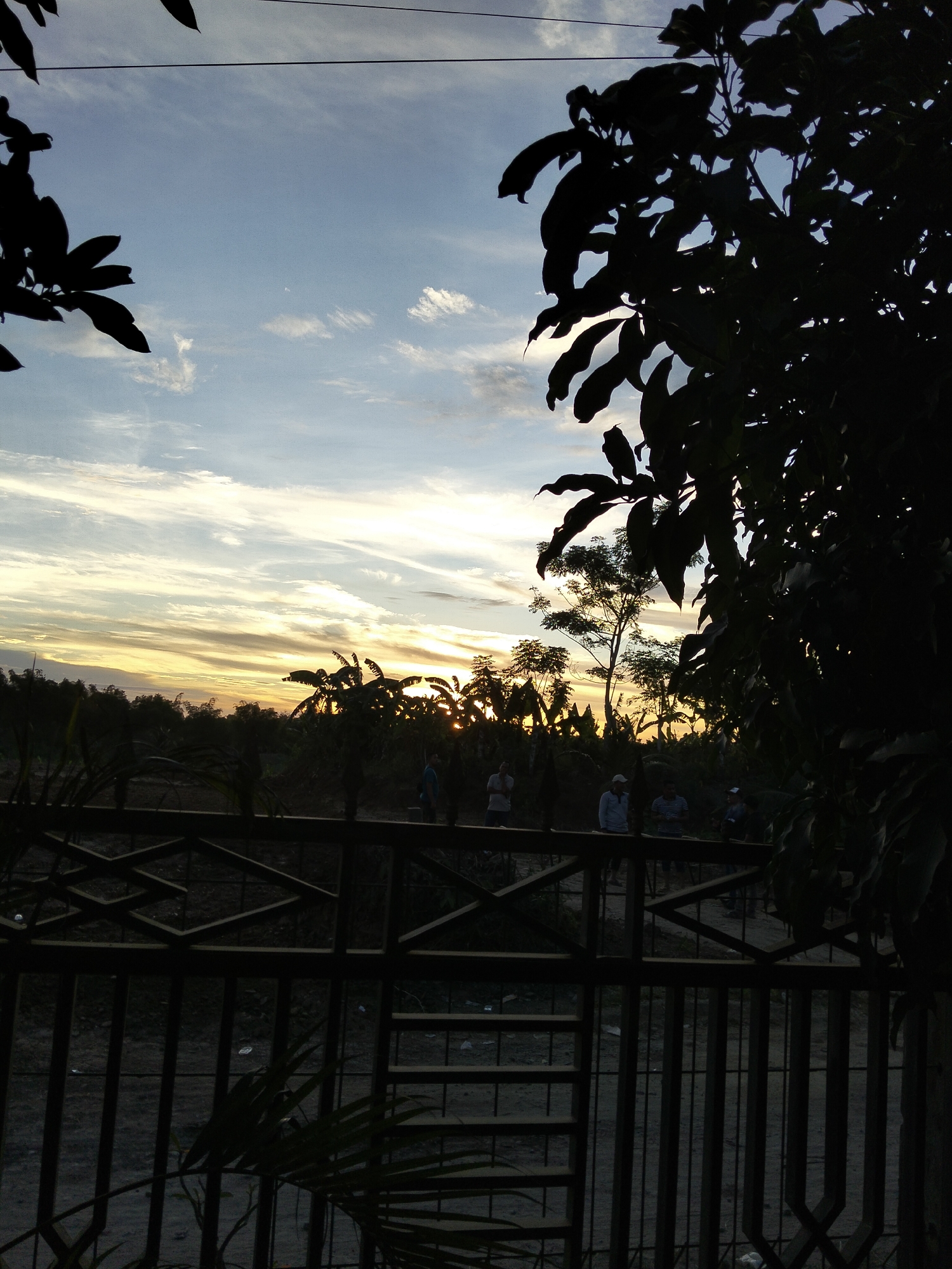 The image depicts a silhouetted scene at dusk or dawn, with a fence and trees in the foreground. The sky is filled with streaks of clouds, illuminated by the setting or rising sun, creating a dramatic backdrop. In the distance, a few figures are visible, appearing as dark shapes against the brighter sky. The overall atmosphere is tranquil, with a sense of quiet observation of the natural light. The location is identified as Payagaboh, Indonesia.
