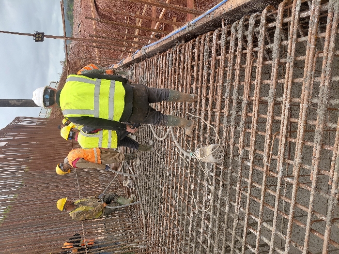 An elevated view shows a construction site in Fdalate, Morocco, where workers are engaged in pouring concrete onto a reinforcement steel mat. Six individuals are visible, all wearing hard hats (yellow or white) and high-visibility vests (yellow or orange), consistent with safety attire. The foreground and midground are dominated by a dense grid of rusty rebar, approximately 20-30 cm deep, which is partially filled with wet, grey concrete.
One worker, positioned on the left-center, wears a yellow hard hat and a yellow hi-vis vest, and is operating a concrete vibrator. The vibrator's motor unit rests on the rebar grid, with its flexible shaft immersed in the fresh concrete to consolidate it. Another worker in an orange hi-vis vest and yellow hard hat stands nearby, observing or assisting.
The individual closest to the camera, with their back turned, wears a white hard hat, a yellow hi-vis vest with reflective strips, dark work pants, and muddy boots, suggesting movement through wet concrete or soil.
In the background to the left, a tall wall of densely packed vertical rebar rises, indicating further structural work. To the right, beyond the active pouring area, reddish-brown excavated earth or an embankment is visible under an overcast sky. A vertical metal pole with a light fixture is present on the far right. The overall environment is muddy and industrial.