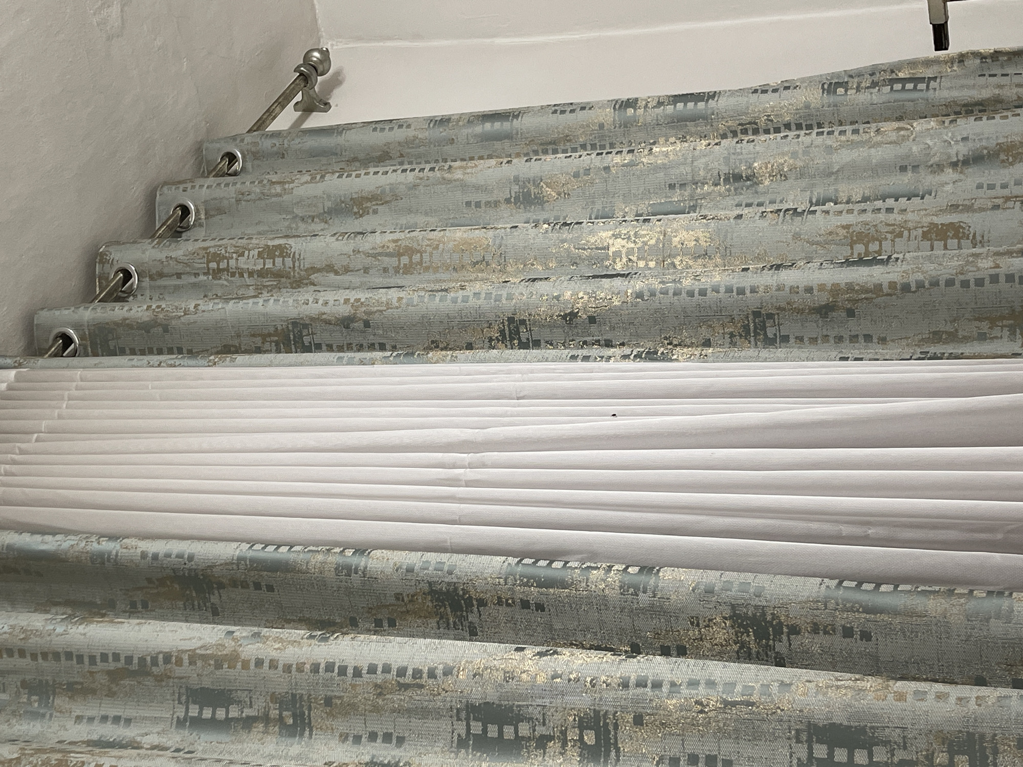 The image shows a close-up of two sets of curtains hanging from a ceiling-mounted curtain rod. The curtain on the left and right are a light blue-grey color with a textured pattern of gold and dark grey squares. The curtain in the middle is a sheer white fabric that appears to be pleated. The curtains are held up by metal grommets that thread through a metal curtain rod. The rod is supported by a metallic bracket mounted to the ceiling. The scene is indoors and appears to be a residential or commercial space. There is no indication of the city of Bishkek, Kyrgyzstan.