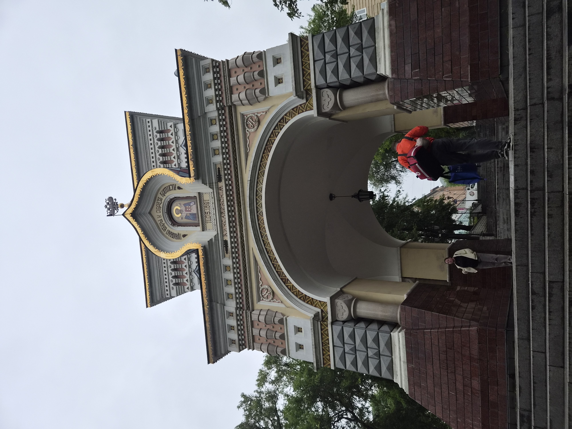 This image captures the Triumphal Arch in Vladivostok, Russia, on an overcast day. The arch, a grand structure with intricate architectural details, is the central focus. It features ornate golden trim, a central icon, and decorative stonework in shades of grey, pink, and beige. The base of the arch is clad in dark reddish-brown stone.
In the foreground, a set of wide stone steps leads up to the arch. Two individuals are visible on these steps. On the left, a woman stands facing the arch, wearing a light-colored jacket and dark pants. Further up the steps, on the right side, a person wearing a bright orange jacket and dark pants is ascending, carrying a large red bag and a blue bag on their back. The person is captured in motion, walking away from the camera and towards the arch.
Behind the arch, glimpses of trees and buildings can be seen, suggesting an urban environment. A black lantern hangs from the ceiling of the arch's interior. The overall atmosphere is subdued due to the overcast sky.
