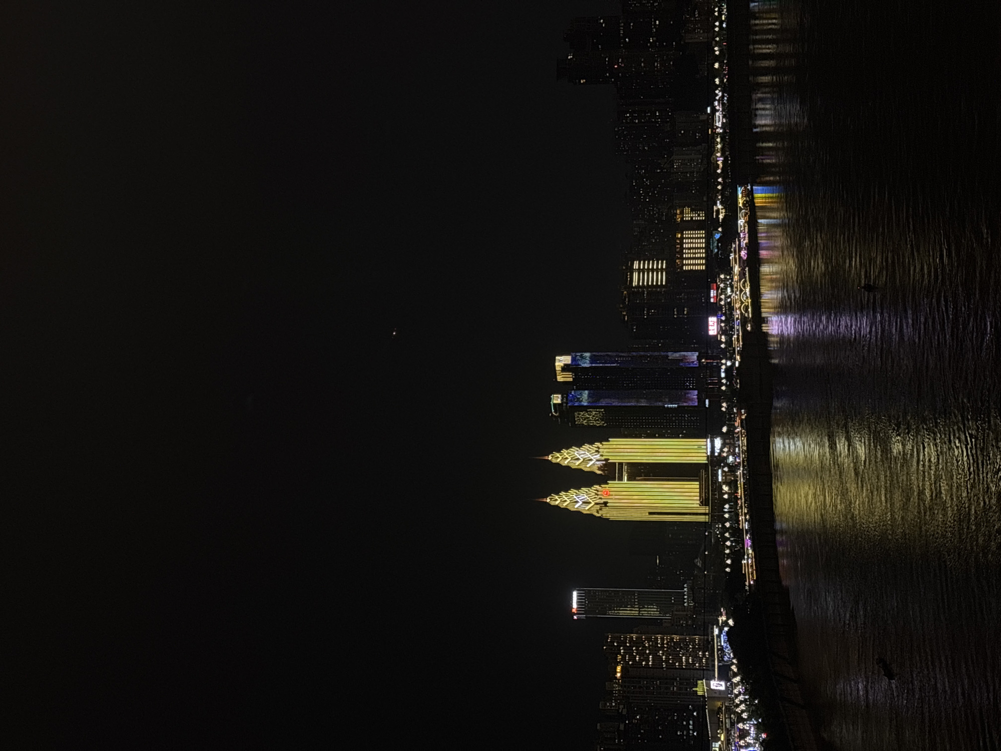 The image captures a vibrant night cityscape along a river, likely taken from a high vantage point in Yuzhong District, China. The scene is dominated by a dark night sky that occupies the left half of the frame, contrasting with the brilliantly lit urban development on the right.
Tall, modern buildings form a dense skyline along the riverbank. Many structures are illuminated with various artificial lights, creating a dazzling display. Two prominent skyscrapers stand out with distinctive golden, geometric lighting patterns that extend up their facades. Other buildings feature patterns of blue, white, and yellow lights. The overall illumination suggests a bustling, modern city.
The wide river stretches across the right side of the image, reflecting the myriad of city lights in shimmering, elongated streaks of color, including yellow, blue, and purple hues. The water surface appears to have gentle ripples, causing the reflections to undulate. A small, dark, indistinct object, possibly a boat or vessel, is visible on the river in the middle-ground, adding a subtle element of activity to the otherwise still waters.
The time of day is clearly night, with no natural light sources visible. The weather appears clear, allowing for unobstructed views of the city lights and their reflections. There is no discernible activity involving people, but the brightly lit city implies constant human presence and urban life. No clearly legible text or signage is visible on any of the buildings or boats. The image conveys a sense of grandeur and the dynamic energy of a major metropolitan area after dark.