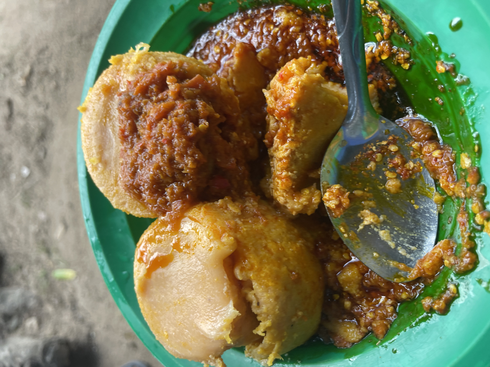 A close-up, eye-level shot captures a green plastic plate filled with a spicy dish, likely an African cuisine from Jos, Nigeria. The plate contains what appear to be dumplings or fritters, coated in a rich, red, and slightly oily chili sauce. Some of the dumplings are whole, while others are cut, revealing a soft, pale interior. A metal spoon rests on the side of the plate, its surface reflecting the dish and partially submerged in the sauce. The background is blurred, showing a rough, sandy surface with some pebbles, consistent with an outdoor setting in Jos. The lighting suggests it is daytime, with the light source likely coming from above and slightly to the left. The overall presentation is rustic and appetizing, emphasizing the texture and vibrant color of the food.