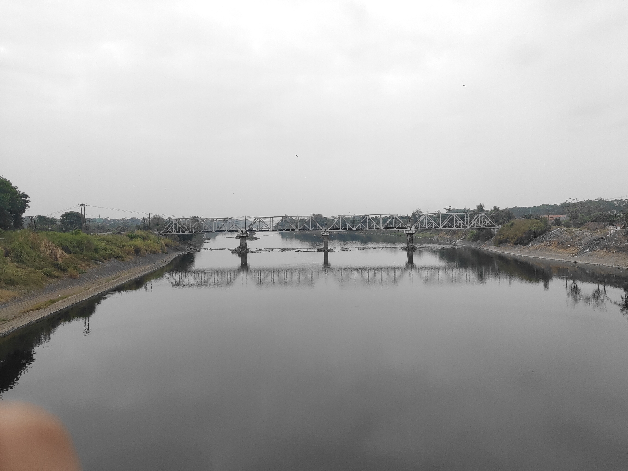 The image shows a wide, dark body of water, likely a river, with a metal truss bridge spanning across it. The bridge has multiple supports and a complex network of beams. The water's surface reflects the bridge and the overcast sky, creating a symmetrical, muted scene. On the left bank of the river, there is a sloping embankment covered in green vegetation and some dry grass. Further back, a few trees and power lines are visible. The right bank appears to be a more barren, rocky slope with some sparse vegetation. In the distance, along the right bank, a few buildings are visible, suggesting a populated area. The sky is a uniform, light gray, indicating a cloudy day. The overall impression is one of a functional, industrial landscape softened by the natural element of the river and the muted lighting.