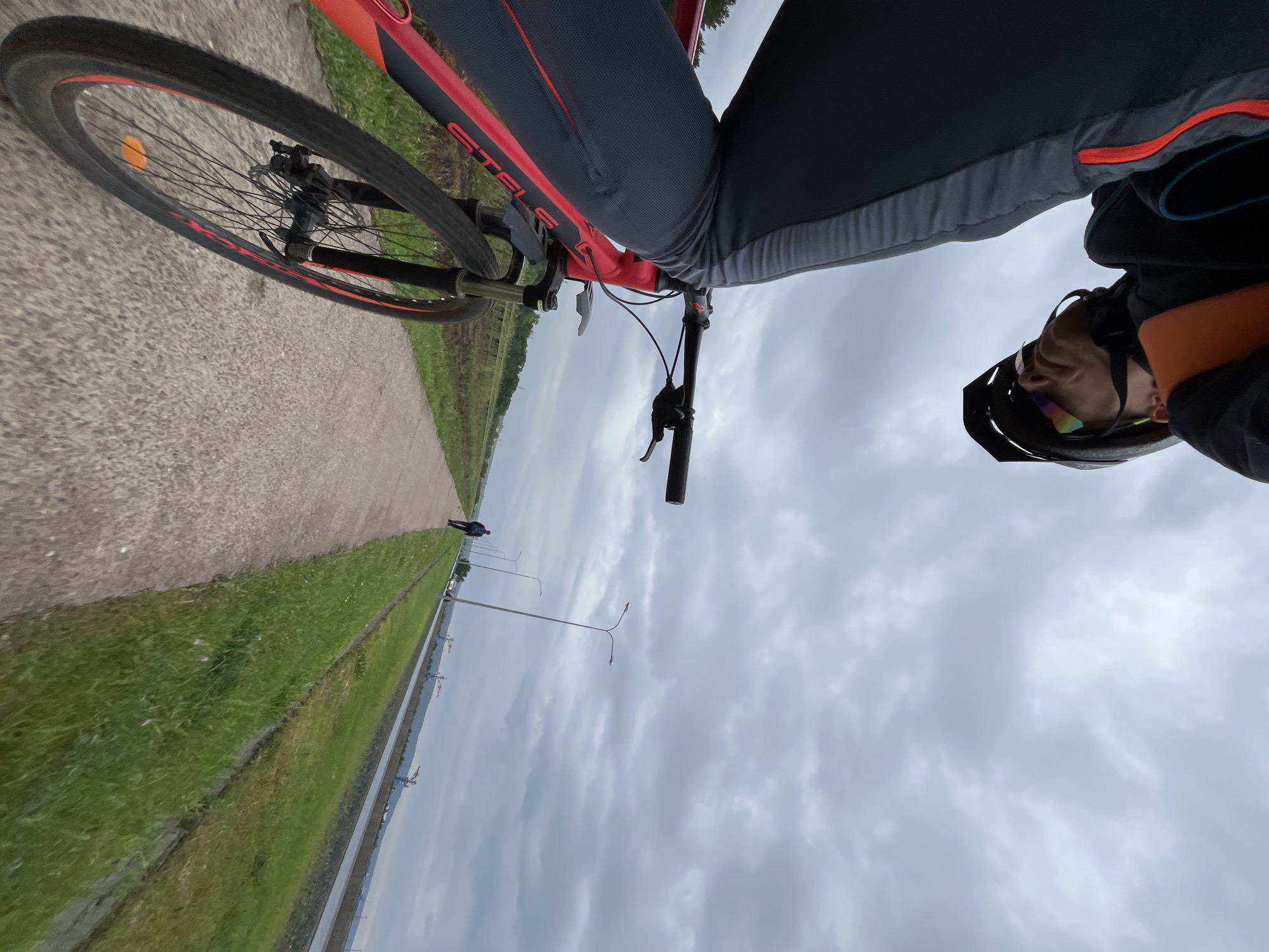 The image provides a tilted, first-person perspective of a person cycling on a paved path in Chaikovsky, Russia, under an overcast sky. The rider's legs, clad in dark grey cycling trousers with red and grey accents, are visible at the center-right. The rider also wears a black helmet with a visor and iridescent sunglasses.
The front portion of a red and black bicycle is prominently featured, with "STELS" written in white on its frame. The handlebars are visible, and the front wheel, equipped with a black tire and red rim accents, is angled towards the left, resting on the paved surface.
The path is bordered on the left by a strip of vibrant green grass, which in turn leads to a calm body of water, likely a canal or river. Across the water, in the distance, what appear to be industrial structures or cranes are faintly visible. Further along the paved path, a lone individual in dark clothing walks away from the camera. Several lampposts line the path, receding into the distance. The sky is dominated by a uniform layer of grey, overcast clouds, suggesting a cloudy day.