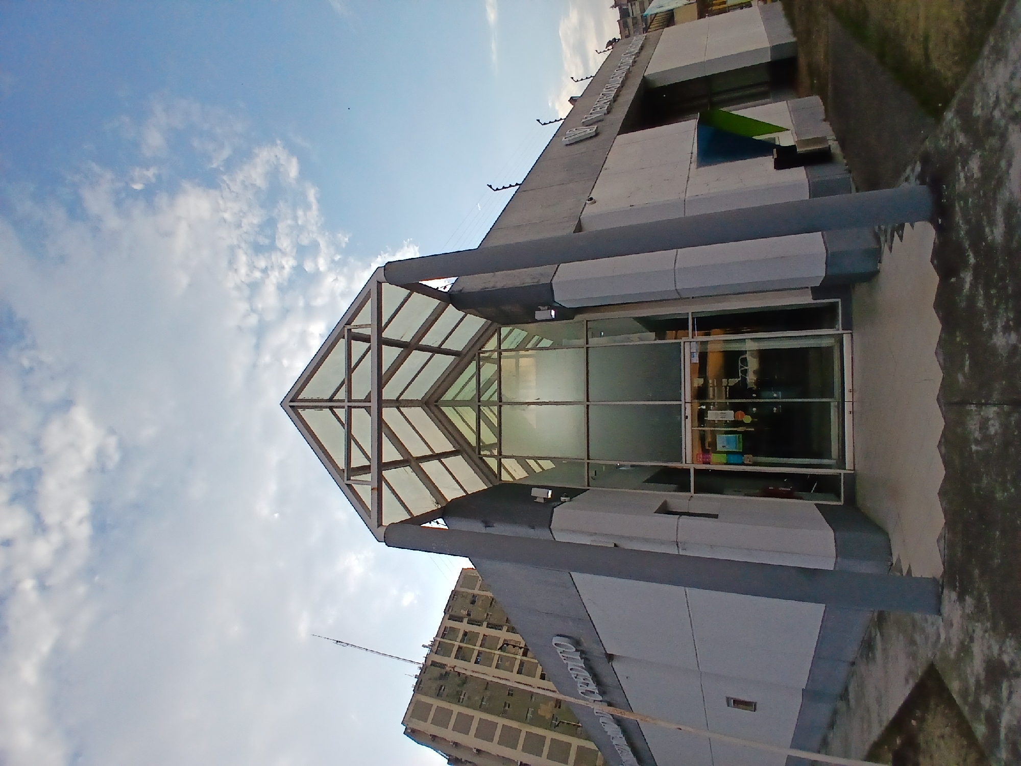 A tilted, daytime photograph captures a modern building, likely a commercial center, against a bright blue sky dotted with scattered white clouds. The primary building features a prominent, angular glass and light-colored metal structure, which appears to be part of a skylight or a grand entrance canopy. Its facade is constructed from alternating sections of light grey and dark grey panels, incorporating large reflective windows.
The image is rotated counter-clockwise, causing the building to appear diagonally across the frame and the foreground ground level to rise sharply on the right. This foreground consists of a textured, possibly concrete, paved area.
On the building's upper facade, large, white, three-dimensional letters are mounted, clearly spelling "Centro Comercial" (Commercial Center). Below some of these letters, small, dark, wavy decorative elements are visible. Further along the building's exterior, a distinct green and blue angular element protrudes, possibly an awning or a decorative architectural feature.
In the background, behind the modern structure, an older, multi-story urban building with numerous windows or balconies is partially visible. A slender antenna or rod extends vertically from behind the buildings into the sky. No individuals or specific activities are depicted, emphasizing the architectural form. The bright daylight and clear sky indicate fair weather, likely in the morning or afternoon in Ciudad Guayana, Venezuela.