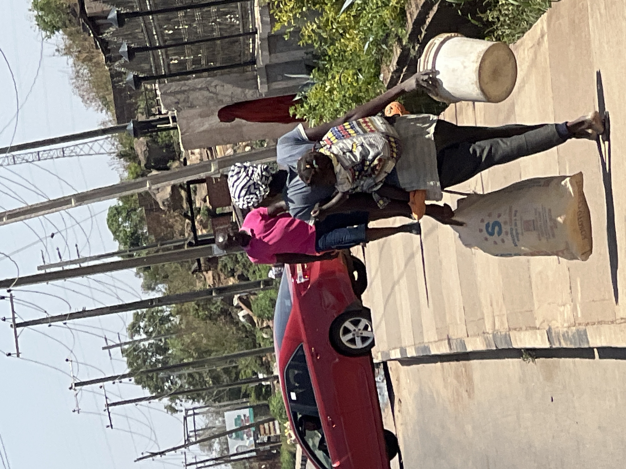In Gura Top, Nigeria, along a paved road lined with utility poles, a man and woman are transporting goods using a pole. The man, wearing a light grey shirt and dark pants, carries a white bucket and a large sack labeled with the letter "S" at either end of the pole. The woman, wearing a pink shirt and blue jeans, assists in carrying the pole. A red car is parked nearby. In the background, there is a building with a fence on top of a rocky formation.