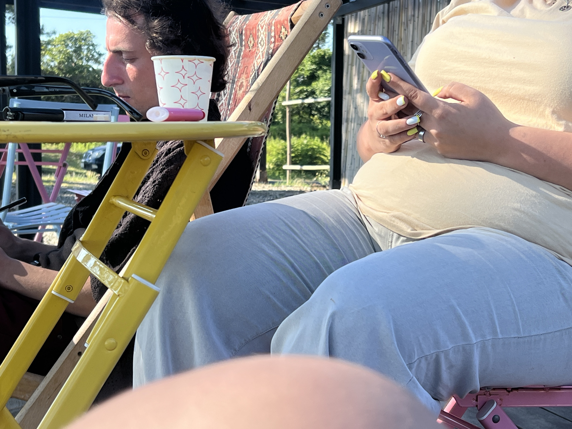 Two people are sitting outdoors on a sunny day, possibly in Ochamchire, Abkhazia. The person on the left, a man with dark, curly hair, is looking down at a yellow table. On the table, there's a white paper cup with red star-like patterns and a pink object that appears to be a vape pen. Next to it are a pair of dark-rimmed glasses and a cigarette pack labeled "MILAN". The man is wearing a dark shirt.
To the right, another person, whose face is not fully visible, is holding a smartphone in their hands and appears to be texting or browsing. This person is wearing a light beige top and light blue pants. Their fingernails are painted yellow and white. They are sitting in a deck chair with a patterned fabric. The background shows lush green trees and bamboo-like structures, suggesting a warm, possibly tropical or subtropical climate. The sun is casting strong shadows, indicating it is likely midday or late afternoon. The overall mood seems relaxed, with both individuals engaged in their own activities.