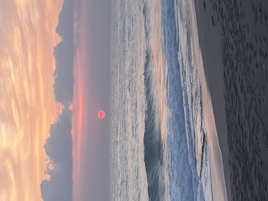 The image displays a sunset over the sea at a beach in Alanya, Turkey. The upper portion of the frame features a sky with horizontal bands of pale orange and pink, transitioning to a softer purple-pink closer to the horizon. Grey-blue cumulus clouds are visible above the horizon. A prominent, spherical, bright red-orange sun is centered just above the sea. The middle section shows the calm surface of the sea, reflecting ambient sky colors, with several lines of white-capped waves breaking and receding towards the sandy shore. The foreground consists of dark, wet sand, marked by faint textural variations. A small, indistinct green item is present on the sand in the lower right corner. No individuals or significant artificial structures are visible within the frame.