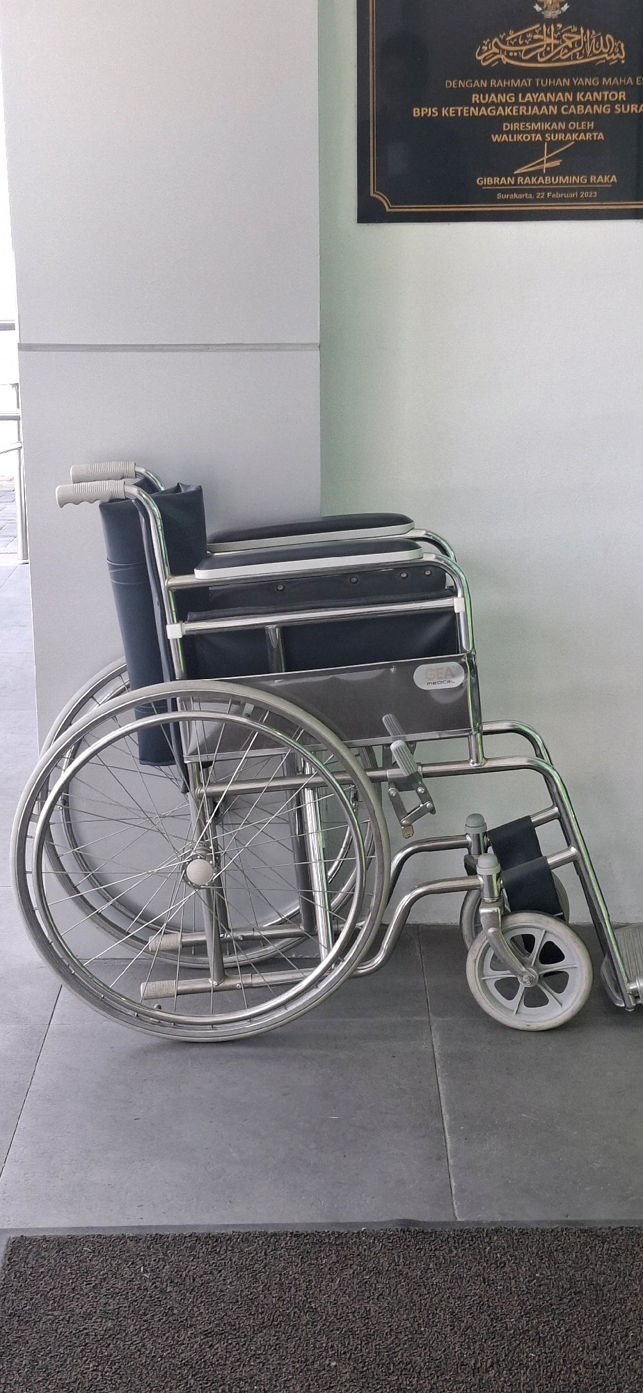 An empty, metallic wheelchair is positioned against a light gray wall. The wheelchair has large spoked wheels with rubber tires, smaller front wheels, and dark blue upholstery for the seat and backrest. Silver push handles are visible at the top of the backrest. A dark brown doormat is in the foreground, and a plaque with Indonesian text is mounted on the wall behind the wheelchair. The plaque indicates the opening of "RUANG LAYANAN KANTOR BPJS KETENAGAKERJAAN CABANG SURAKARTA" (Office Service Room of BPJS Ketenagakerjaan Surakarta Branch) by the Mayor of Surakarta, Gibran Rakabuming Raka, on February 22, 2023.