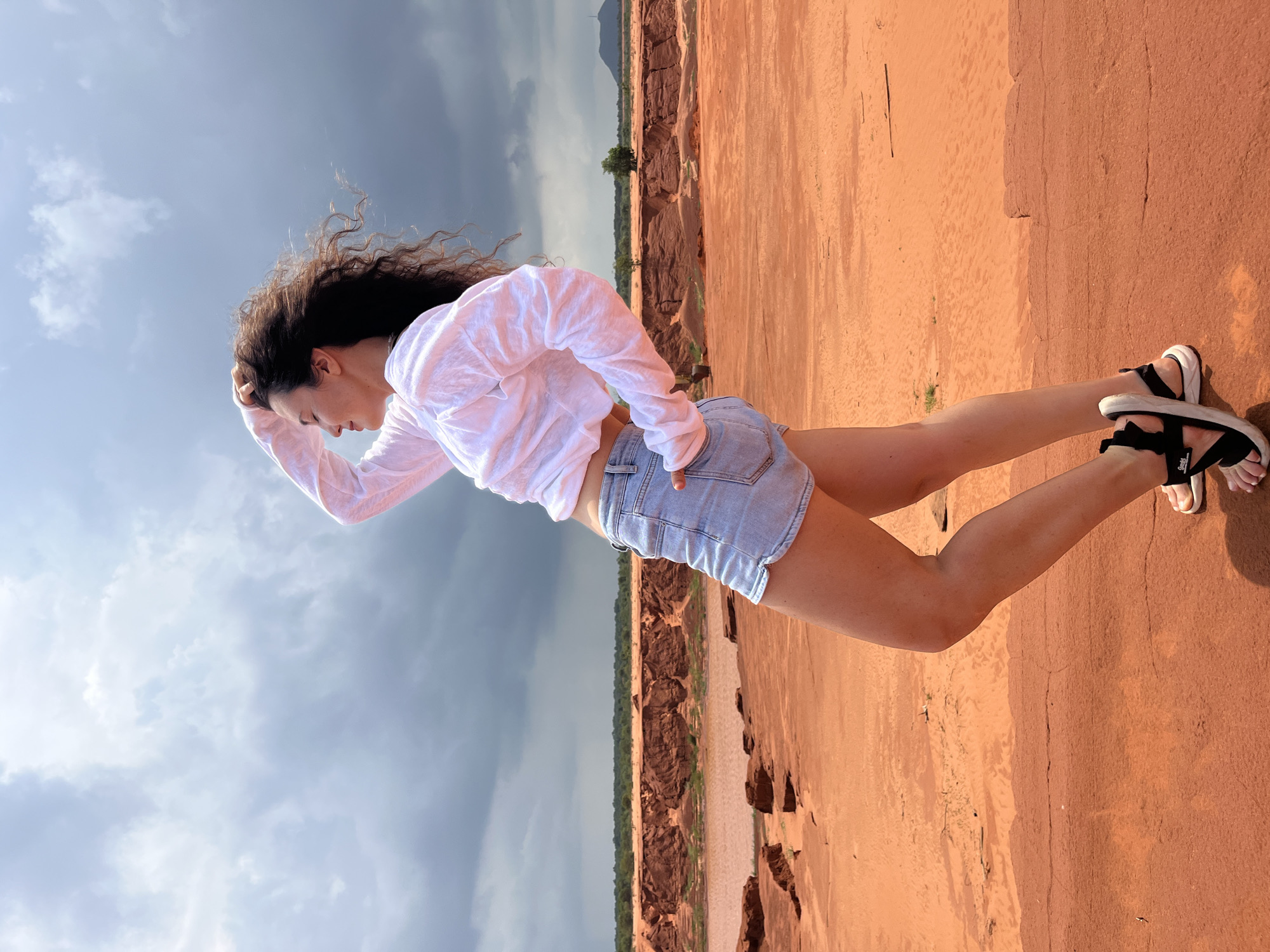 A woman with curly dark hair stands in profile, facing left with her eyes closed and her right hand in her hair, against a dramatic cloudy sky. She wears a white, loose-fitting long-sleeved shirt tucked into light-wash denim shorts. Her left hand is tucked into the back pocket of her shorts. She wears black and white sandals. The woman is positioned on a reddish-orange sandy surface. In the background, there is a body of water, eroded reddish-brown cliffs, and a distant green treeline under a dark, overcast sky. The scene is likely set in the Mui Ne sand dunes, a popular tourist destination in Vietnam known for its picturesque desert-like landscapes.