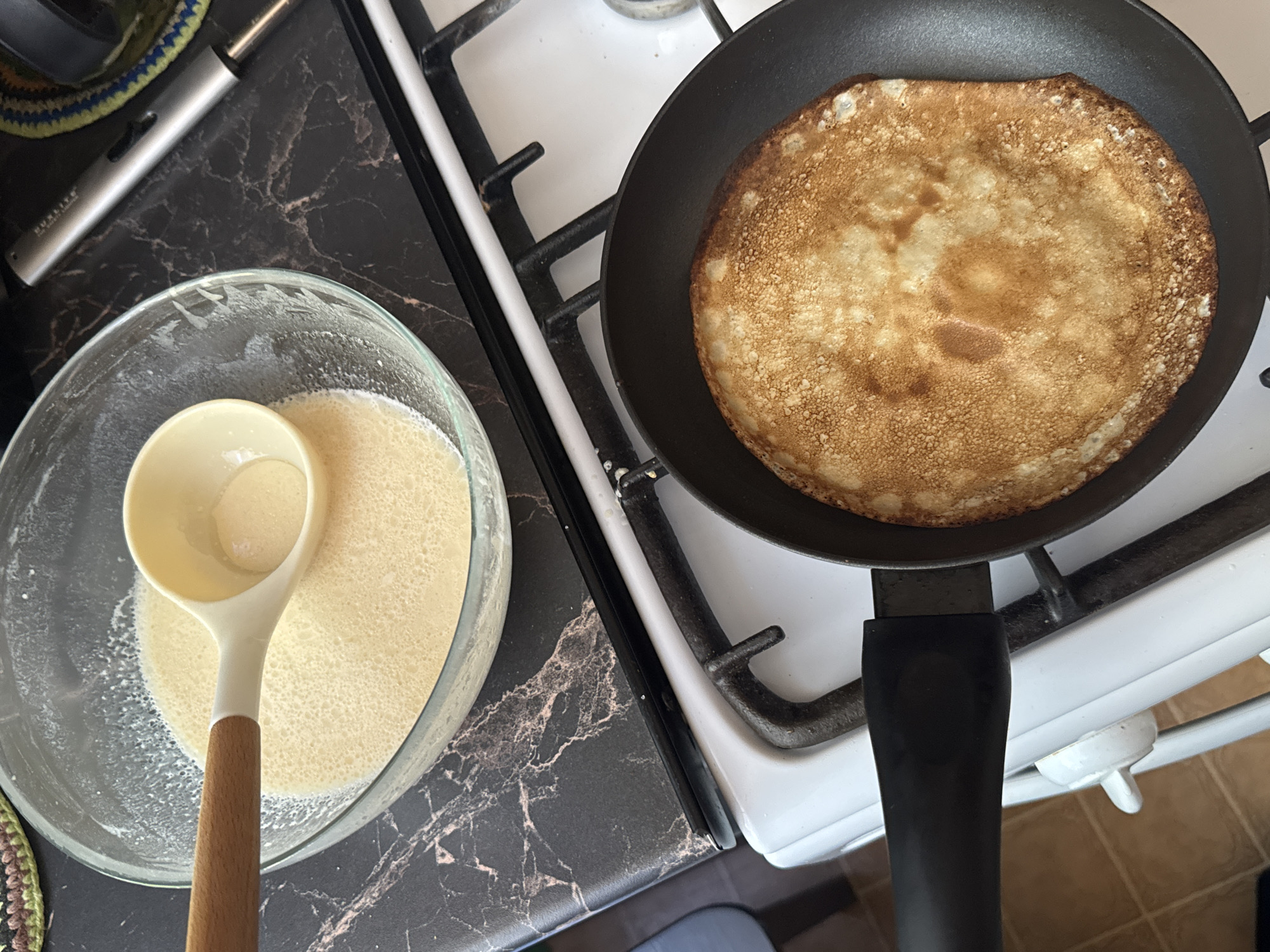 A close-up, overhead shot shows a person preparing crepes in a kitchen in Chaikovsky, Russia. A golden-brown crepe is cooking in a black frying pan on a white stovetop. Next to the pan, a clear glass bowl contains a pale yellow batter, with a ladle resting inside. The countertop is dark marble, and a silver lighter is visible to the right of the bowl. The scene suggests a domestic setting, possibly a home kitchen, with the focus on the cooking process.