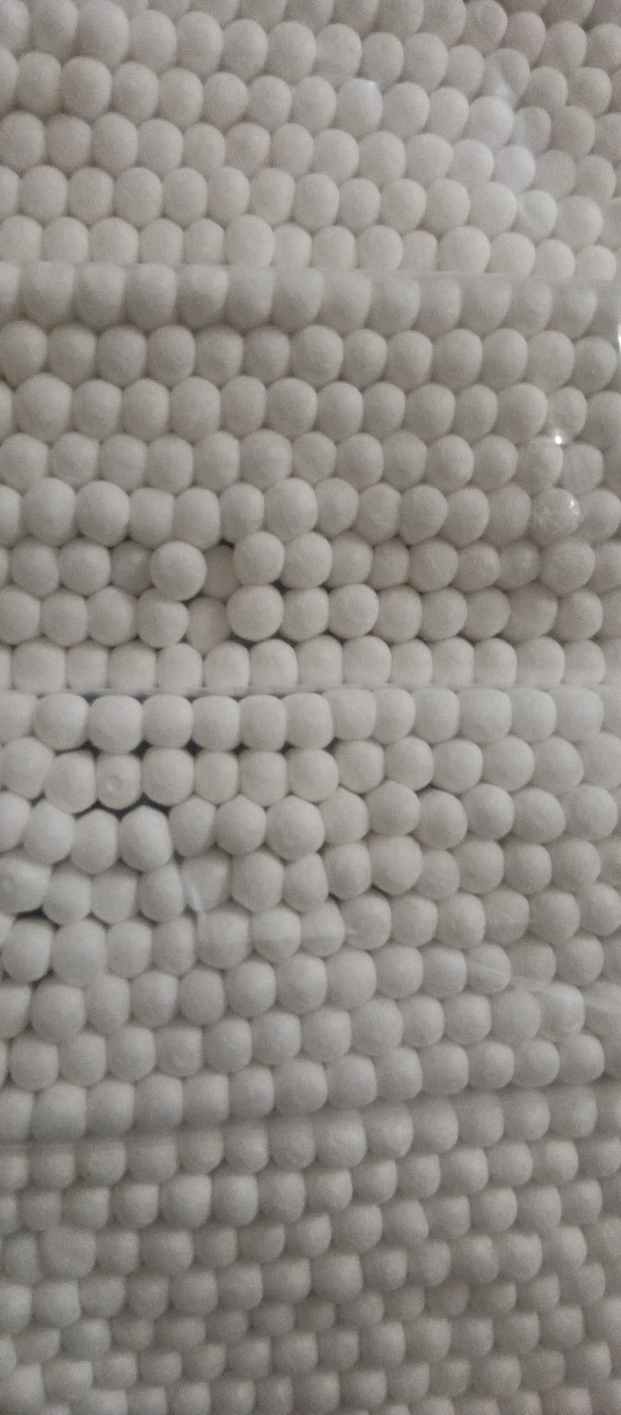 A close-up shows a dense packing of white cotton balls arranged in rows and columns. The cotton balls appear soft and fluffy, filling the frame with their uniform texture. The lighting is soft, casting subtle shadows that accentuate the rounded shapes of the cotton balls. The environment is undefined, with the focus solely on the material and its arrangement. The location is unknown.
