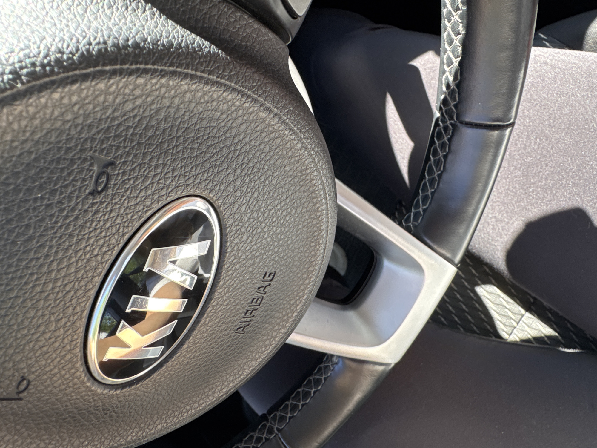 The image shows a close-up of a Kia steering wheel, specifically the emblem and the "AIRBAG" inscription. The steering wheel is made of black leather with white stitching visible on the lower half. The Kia logo is silver and oval-shaped. The horn symbol is also visible on the upper right side of the steering wheel. Sunlight is casting shadows on the steering wheel and the interior of the car. The image is taken from inside the vehicle, looking towards the driver's side. There are no people visible in the image. The scene appears to be within a parked car, likely during daytime. The specific location in Saint Petersburg, Russia cannot be determined from the provided image.