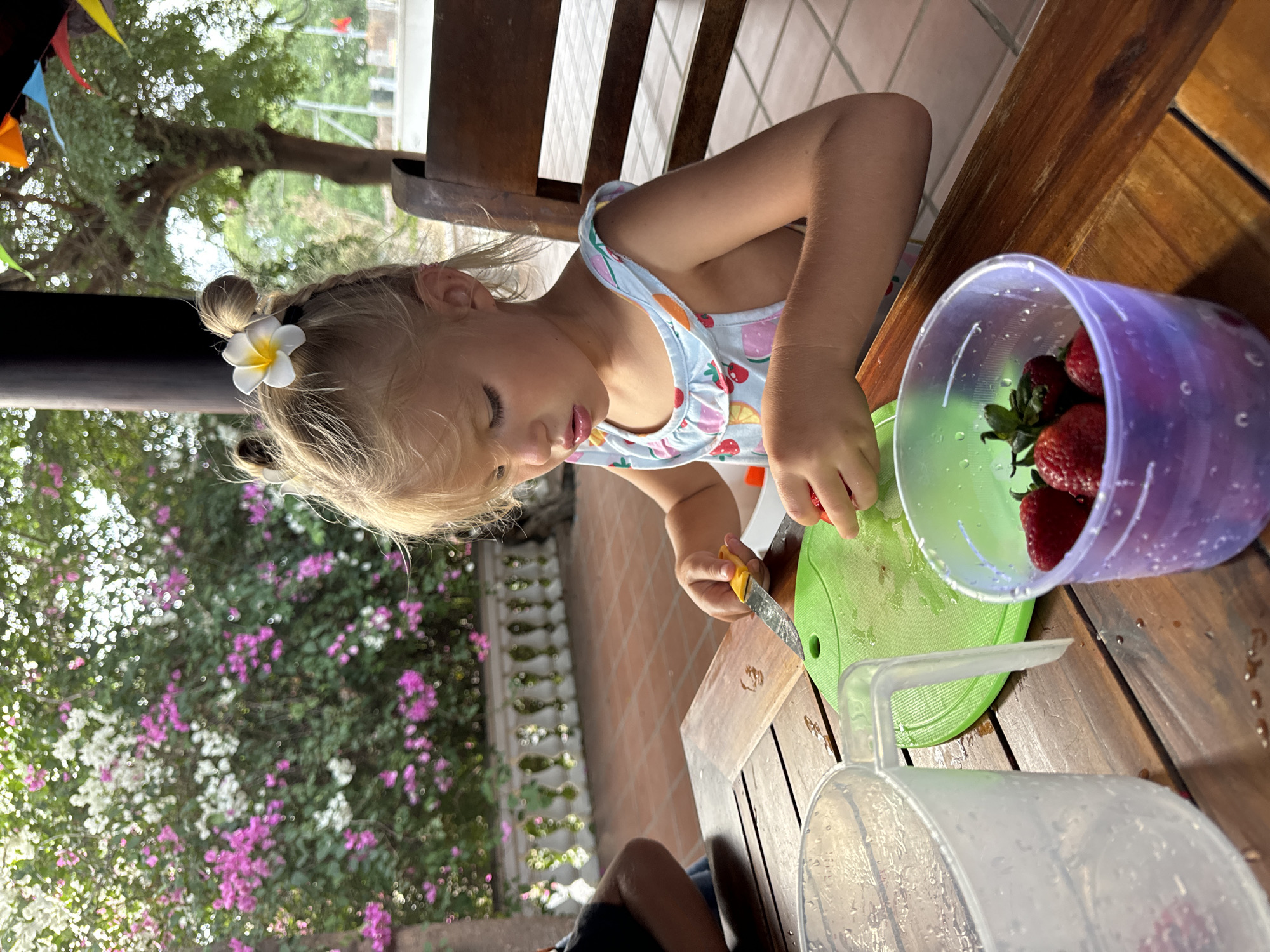 A young girl is sitting at a wooden table outside, possibly in a residential area of Mũi Né, Vietnam, given the tropical foliage and architecture. The girl is blonde and has her hair in pigtails, adorned with a white flower. She is wearing a light-colored tank top with a fruit pattern. She is focused on cutting strawberries with a small knife on a green cutting board. A purple plastic container filled with strawberries is next to the cutting board. Nearby is a clear plastic pitcher or container. In the background is a wall with green foliage and pink and white flowers. Bunting is hung from the roof.