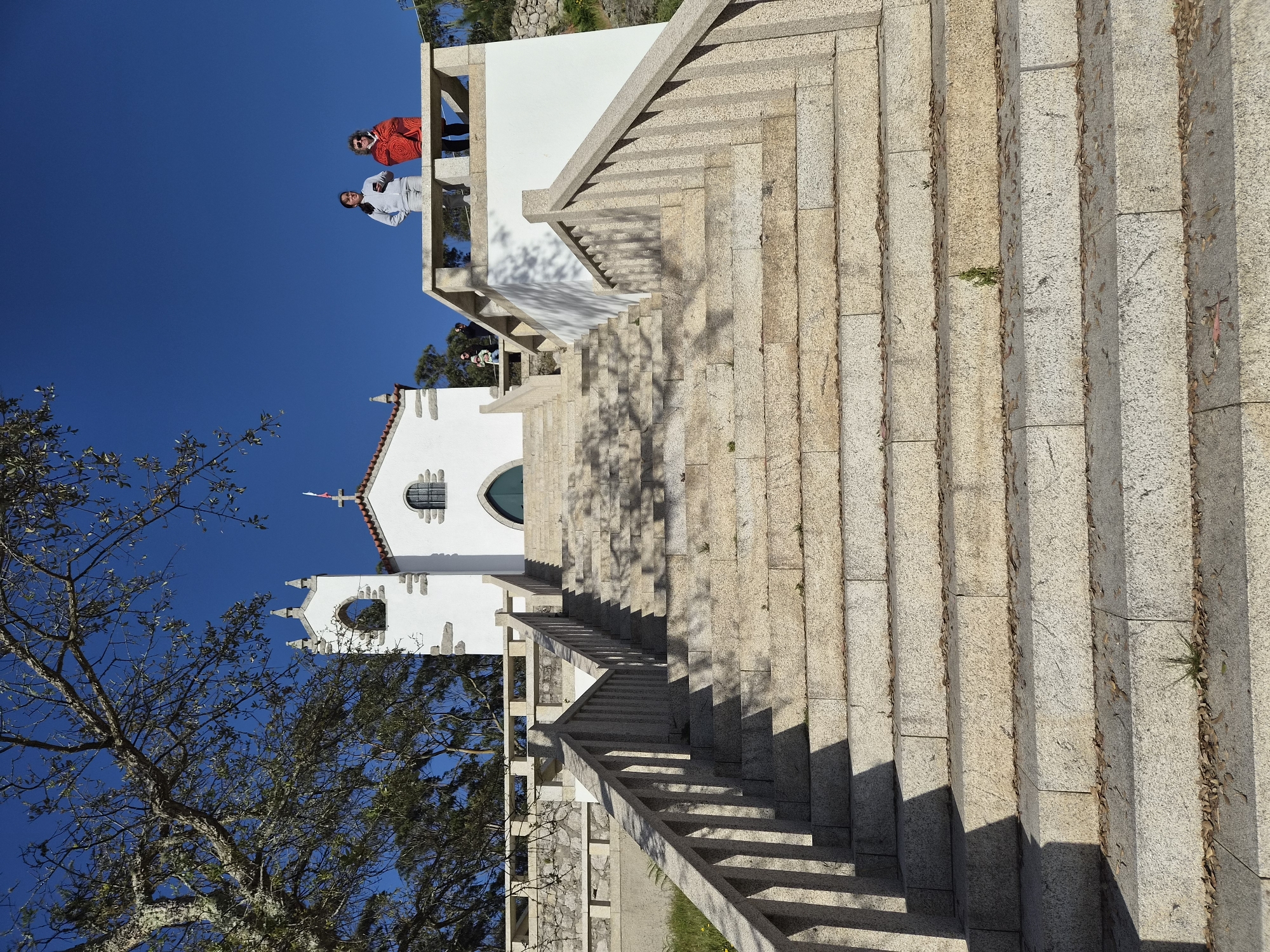 The image shows a long, broad stone staircase leading up to a white church building under a clear blue sky. Two people are standing on an upper balcony. A tree with bare branches is visible to the side of the church. The scene is located in Esposende, Portugal.