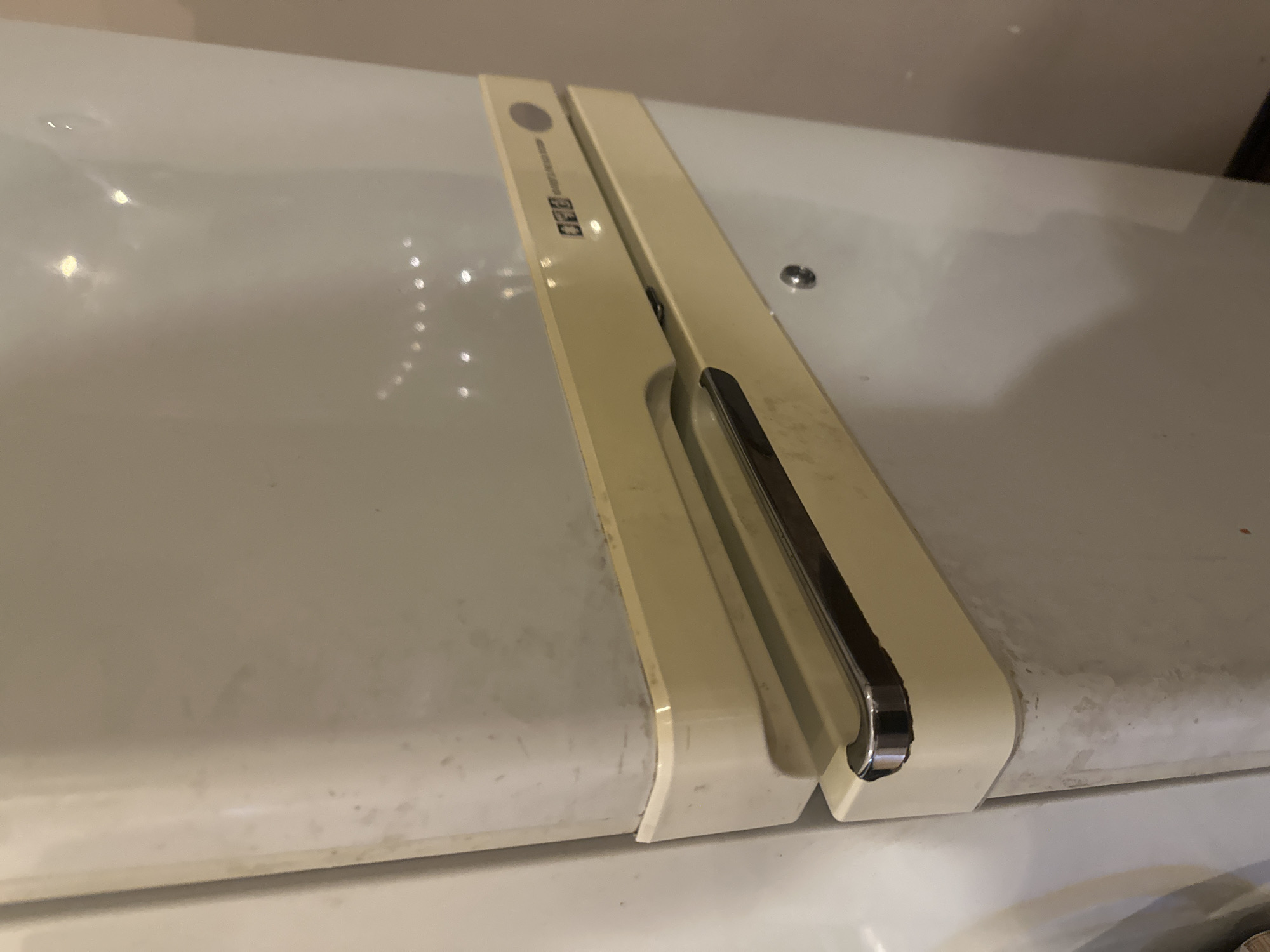 The image shows a close-up view of the cream-colored door of a refrigerator. The refrigerator has a retro design with a textured surface and a horizontal handle with a chrome accent. There are some smudges and marks on the refrigerator door, suggesting it is not new. A silver circular button or dial is visible on the upper part of the door, next to a small black rectangular logo with text. The background is out of focus, but appears to be a wall. The image is not a scene from the city of Orekhovka, Russia.