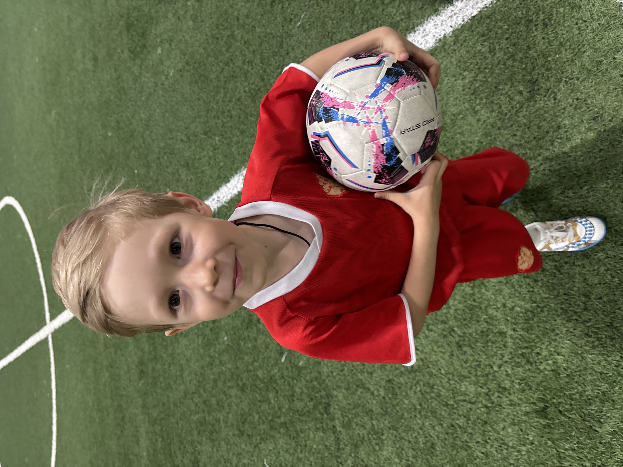 A young boy, likely between the ages of 5 and 7, is holding a "Pro Star" soccer ball. He is wearing a red soccer uniform with white trim. The setting appears to be an indoor soccer field, as evidenced by the artificial turf and white lines on the ground. The location is presumed to be Chaikovsky, Russia.