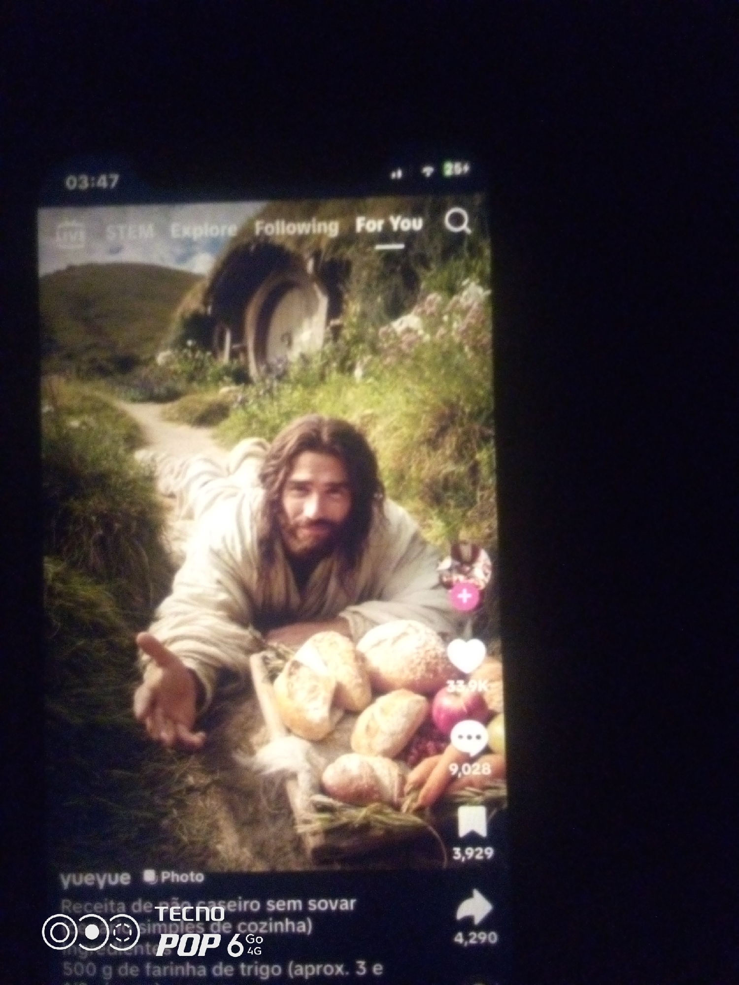The image displays a screenshot of a mobile application, likely a social media platform, featuring a video or image of a man resembling Jesus Christ lying on his stomach in a grassy area. He is wearing a light-colored robe and has long brown hair and a beard. His right arm is extended towards the viewer. In front of him are various baked goods, including loaves of bread and what appear to be fruits like apples and carrots. The background shows a small, round doorway into a hill, reminiscent of a hobbit hole from "The Lord of the Rings," with greenery surrounding it.
On the top left of the screen, the time "03:47" is visible. Along the top bar, there are icons and text indicating "LIVE," "STEM," "Explore," "Following," and "For You," along with a search icon.
The bottom portion of the screenshot contains text related to the video, including a username "yueyue," the word "Photo," and a description in Portuguese: "Receita de pão caseiro sem sovar (simples de cozinha)" which translates to "Recipe for no-knead homemade bread (simple kitchen)." Below this, there is text indicating ingredients, including "500 g de farinha de trigo (aprox. 3 e".
On the right side of the video frame, there are social media interaction icons: a heart with "33.9K" likes, a speech bubble with "9,028" comments, and a bookmark icon with "3,929" shares. Below that is a share arrow icon with "4,290" interactions.
The image is a photograph of a phone screen, and the overall quality is somewhat blurry and grainy. There is no discernible information in the image that indicates the location within the city of Howayi, Nigeria.